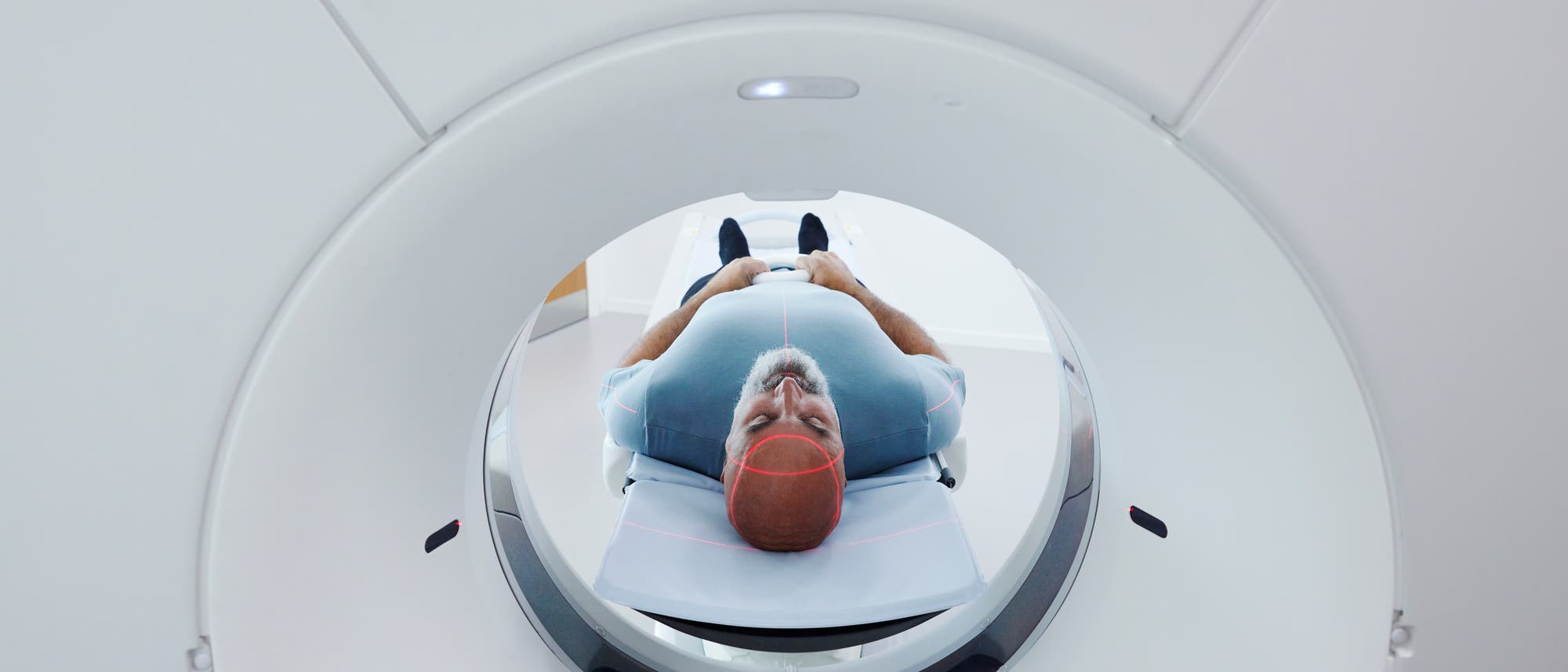 Eine Person liegt auf dem Rücken in einem CT-Scanner. Der Kopf ist in der Mitte des Scanners positioniert, und rote Laserlinien sind auf dem Kopf sichtbar, um die Ausrichtung zu gewährleisten. Die Umgebung ist klinisch und modern gestaltet.