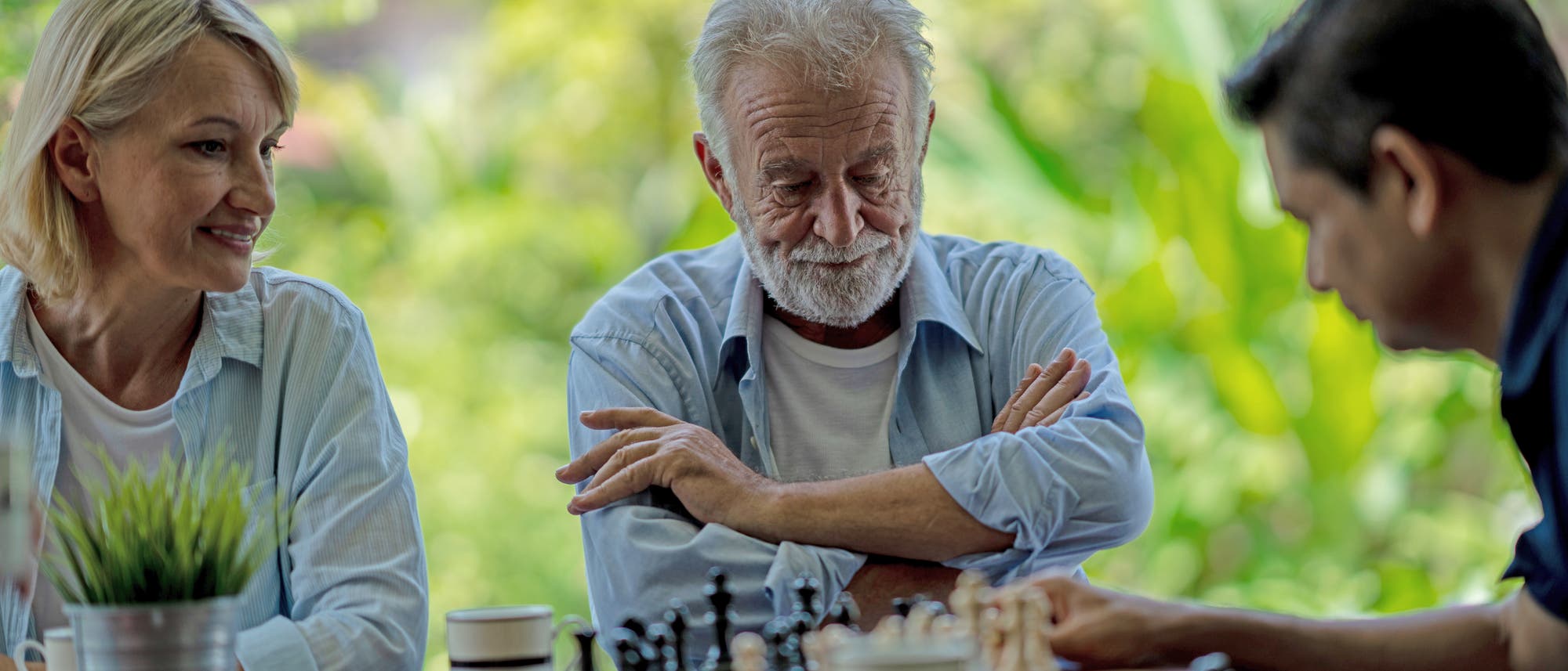 Drei Personen sitzen an einem Tisch im Freien und spielen Schach. Eine ältere Frau und ein älterer Mann beobachten konzentriert das Spiel, während ein jüngerer Mann einen Zug macht. Im Vordergrund steht eine Topfpflanze auf dem Tisch. Der Hintergrund ist grün und verschwommen, was auf einen Garten oder Park hindeutet. Drei Personen sitzen an einem Tisch im Freien und spielen Schach. Eine ältere Frau und ein älterer Mann beobachten konzentriert das Spiel, während ein jüngerer Mann einen Zug macht. Im Vordergrund steht eine Topfpflanze auf dem Tisch. Der Hintergrund ist grün und verschwommen, was auf einen Garten oder Park hindeutet.