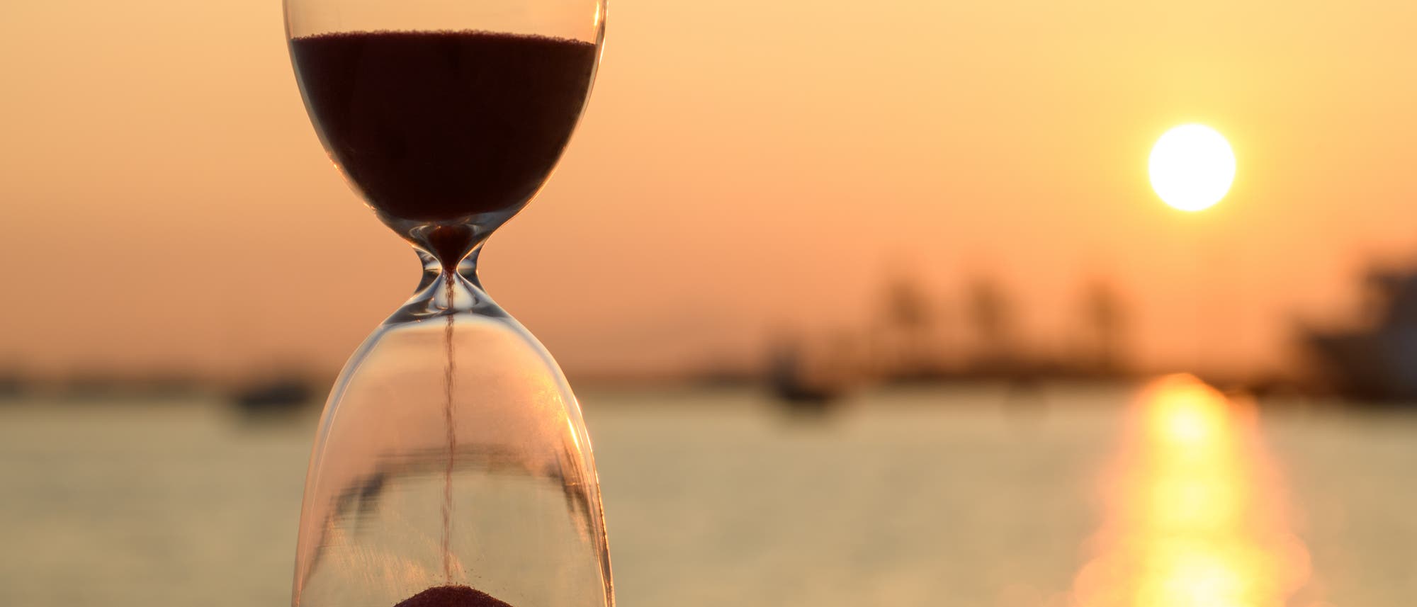 Eine Sanduhr steht im Vordergrund vor einem Sonnenuntergang am Meer. Der Himmel ist in warmen Oranget&ouml;nen gef&auml;rbt, und die Sonne spiegelt sich im Wasser. Die Sanduhr ist halb gef&uuml;llt, und der Sand rieselt langsam von der oberen in die untere Kammer. Im Hintergrund sind verschwommene Umrisse von Booten und einer K&uuml;stenlinie zu sehen.