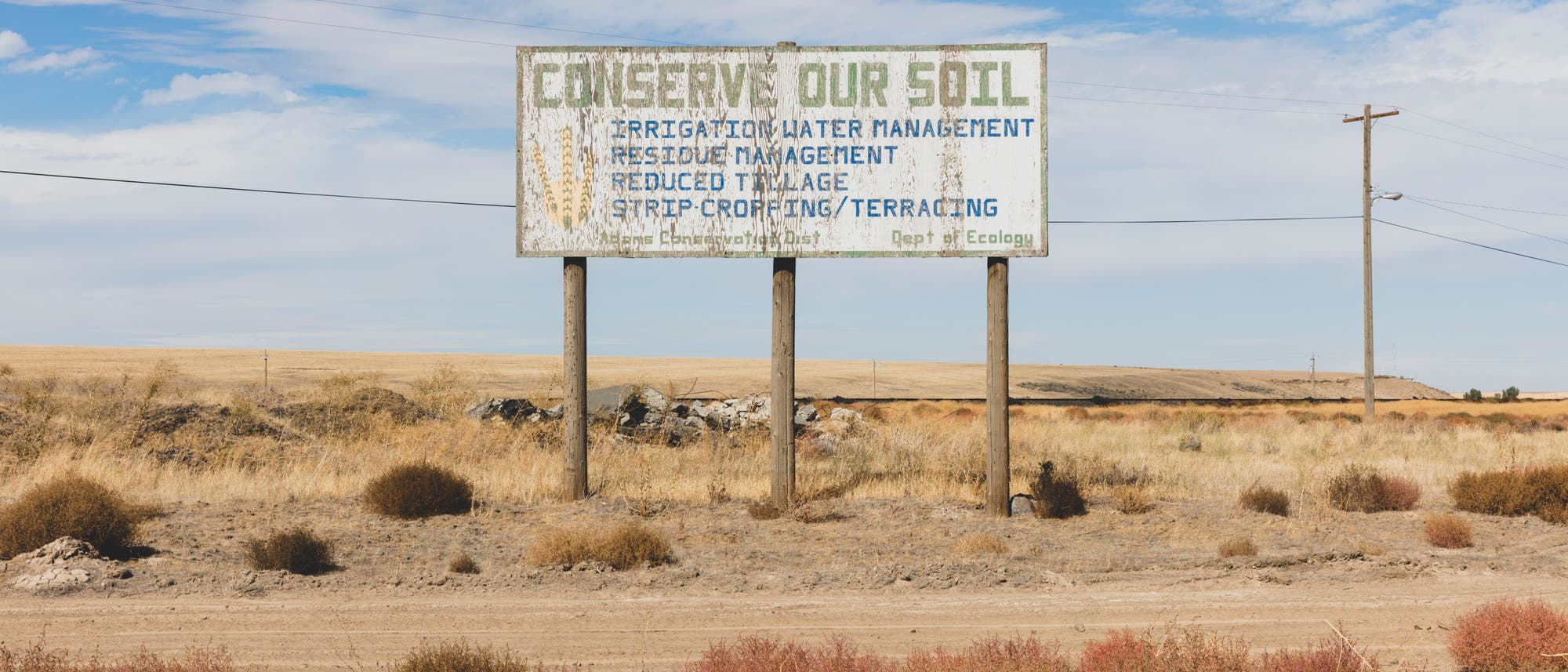 Eine Tafel in einer trockenen Landschaft mit der Aufschrift &bdquo;Conserve Our Soil&ldquo; und weiteren Hinweisen zu Bew&auml;sserungsmanagement, Reststoffmanagement, reduzierter Bodenbearbeitung und Terrassierung. Die Umgebung zeigt eine karge, trockene Landschaft mit vereinzeltem Gestr&uuml;pp und einem klaren Himmel. Die Tafel hebt die Bedeutung des Bodenschutzes hervor.