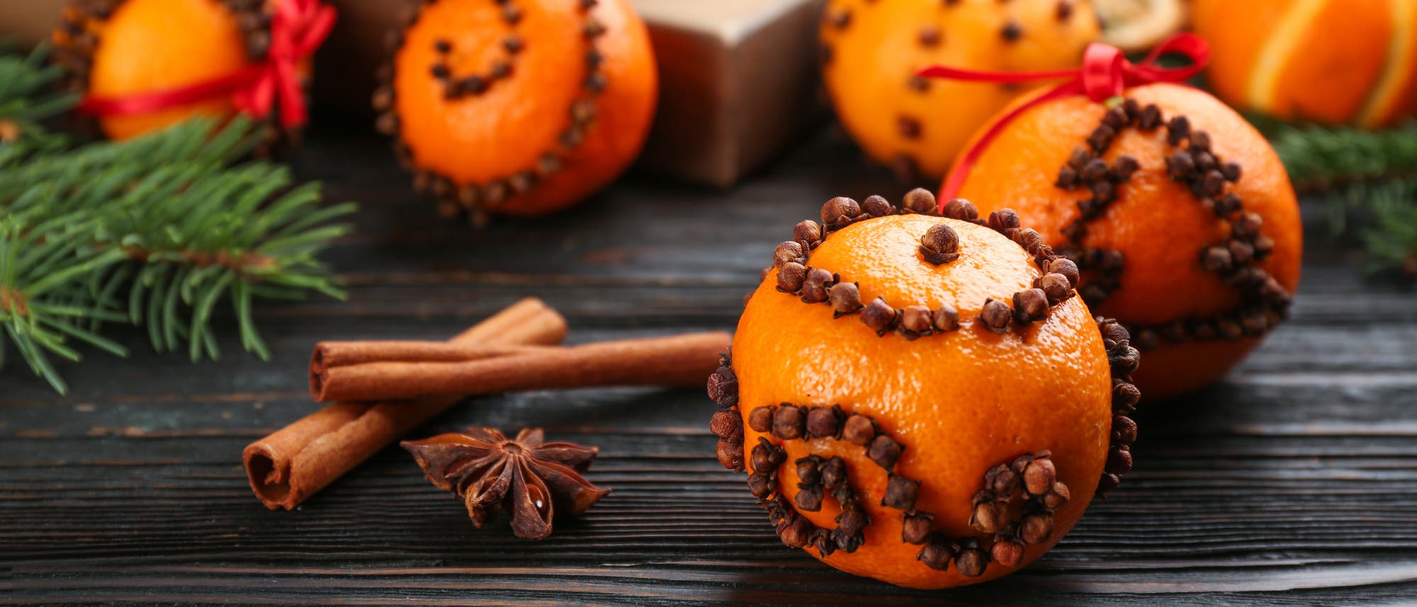 Orangen, die mit Nelken verziert sind, liegen auf einem dunklen Holztisch. Im Hintergrund sind weitere dekorierte Orangen und ein Geschenk zu sehen. Auf dem Tisch befinden sich auch Zimtstangen, Sternanis und Tannenzweige, die eine festliche Atmosph&auml;re schaffen.