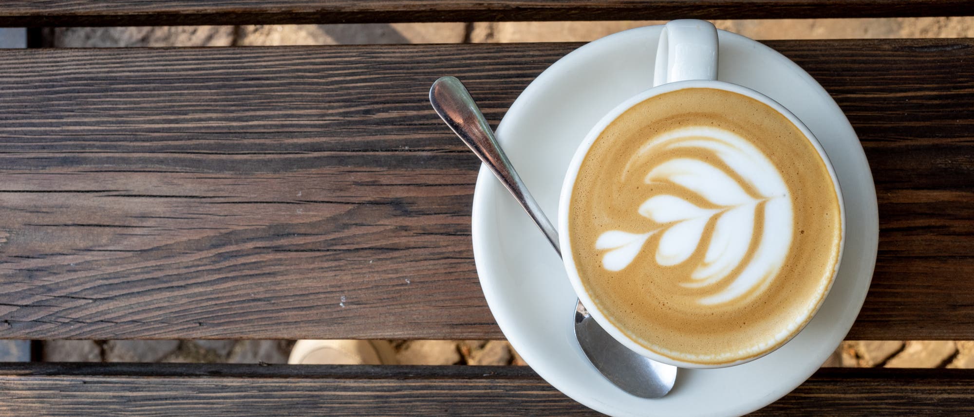 Eine Tasse Cappuccino mit Latte Art in Form eines Blattes auf einem wei&szlig;en Unterteller, daneben ein L&ouml;ffel. Die Tasse steht auf einem Holztisch mit sichtbarer Maserung.