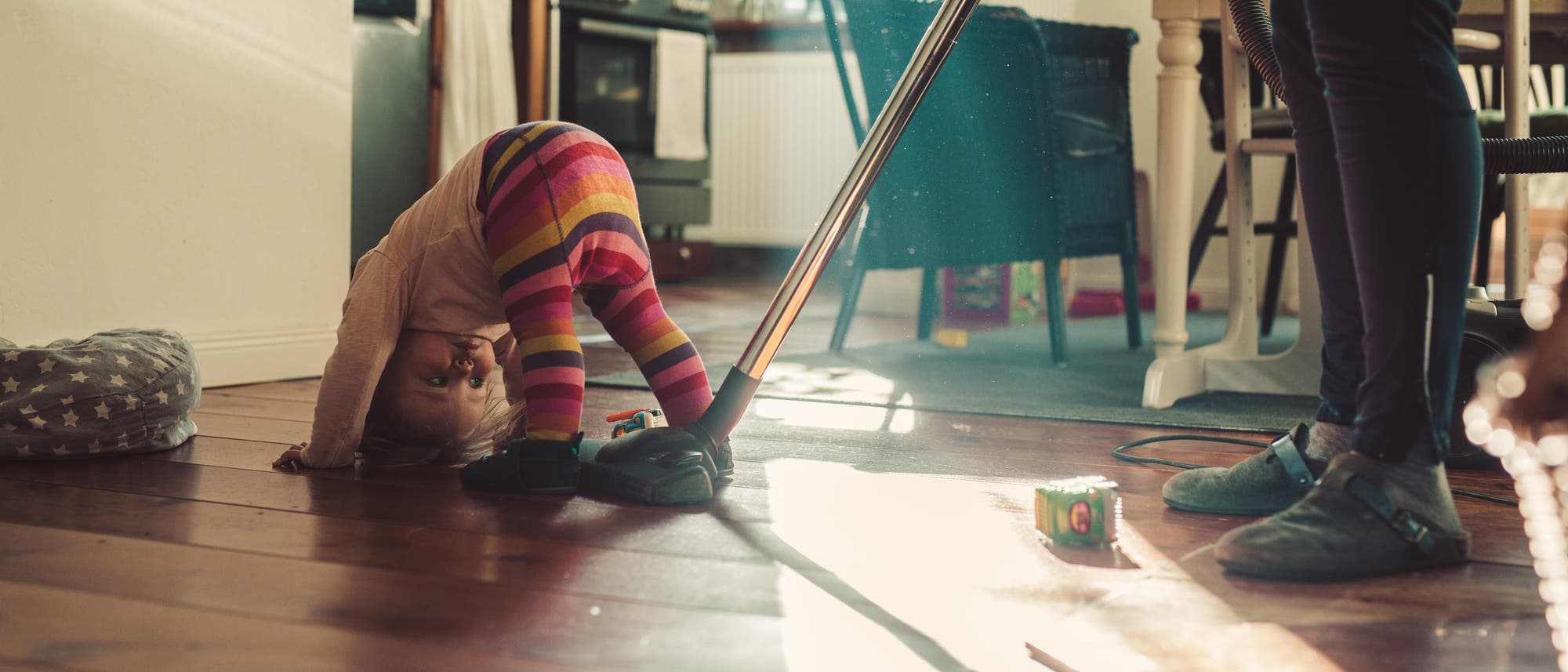 Ein Kleinkind in bunten Streifenleggings spielt auf dem Holzboden eines Wohnzimmers, während eine Person mit einem Staubsauger den Boden reinigt. Im Hintergrund sind eine Küche und ein Esstisch zu sehen. Sonnenlicht fällt durch ein Fenster und beleuchtet den Raum. Ein Spielzeugwürfel liegt auf dem Boden. Ein Kleinkind in bunten Streifenleggings spielt auf dem Holzboden eines Wohnzimmers, während eine Person mit einem Staubsauger den Boden reinigt. Im Hintergrund sind eine Küche und ein Esstisch zu sehen. Sonnenlicht fällt durch ein Fenster und beleuchtet den Raum. Ein Spielzeugwürfel liegt auf dem Boden.