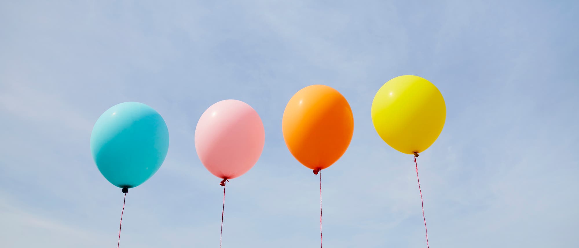 Vier bunte Luftballons schweben an Schn&uuml;ren vor einem blauen Himmel. Die Ballons haben die Farben Blau, Rosa, Orange und Gelb. Sie sind gleichm&auml;&szlig;ig verteilt und vermitteln ein Gef&uuml;hl von Leichtigkeit und Freude.