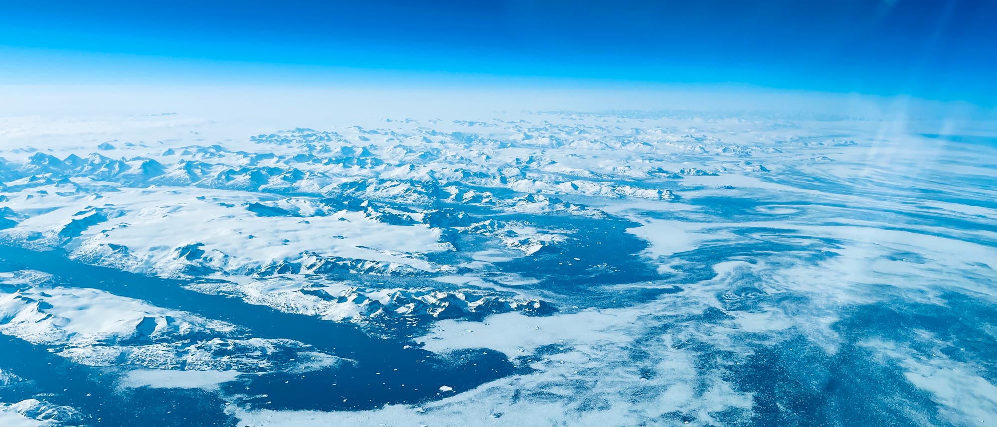 Luftaufnahme einer verschneiten Landschaft mit Bergen und gefrorenen Gew&auml;ssern. Der Himmel ist klar und blau, w&auml;hrend die schneebedeckten Berge und das Eis darunter in verschiedenen Wei&szlig;- und Blaut&ouml;nen schimmern. Die Szene vermittelt eine ruhige, kalte Atmosph&auml;re und zeigt die Weite der unber&uuml;hrten Natur.