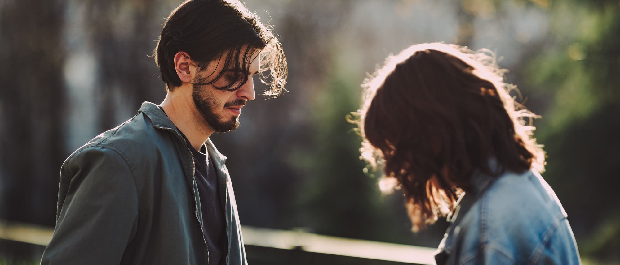 Zwei junge Menschen stehen in einem Park. Die Person links blickt nach unten, während die Person rechts leicht nach vorne geneigt ist. Beide tragen lässige Jacken. Die Szene ist von einem warmen Sonnenlicht durchflutet, das die Umgebung und die Personen sanft beleuchtet. Im Hintergrund sind unscharf Bäume und ein Geländer zu sehen.