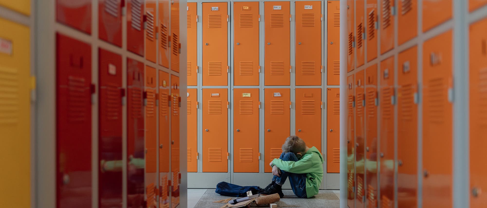 Ein Kind sitzt allein auf dem Boden eines Flurs mit orangefarbenen Schließfächern. Es hat die Knie angezogen und den Kopf darauf gelegt. Neben ihm liegen verstreute Bücher und eine Tasche. Die Szene vermittelt ein Gefühl von Einsamkeit und Nachdenklichkeit.