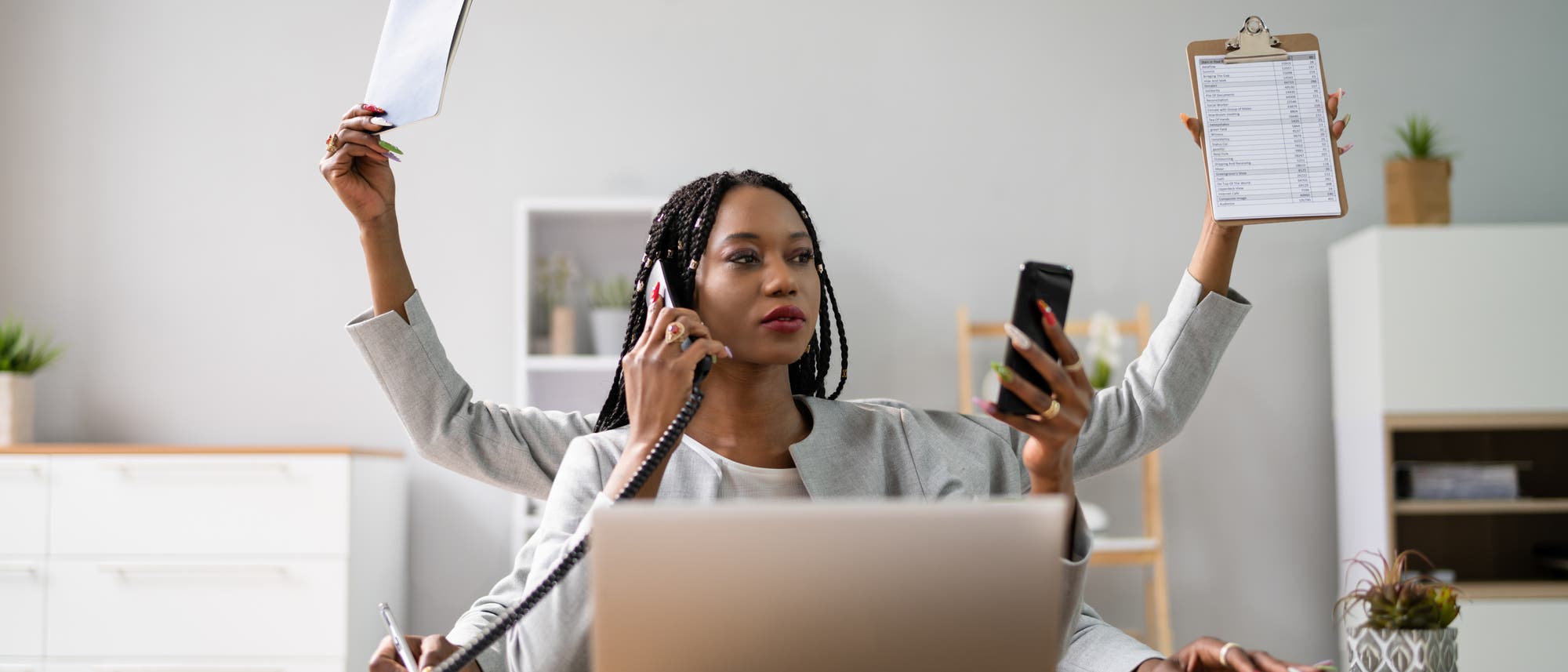 Eine Person sitzt an einem Schreibtisch mit einem Laptop und mehreren Telefonen. Sie h&auml;lt ein Telefon am Ohr, ein weiteres in der Hand und hat zus&auml;tzlich ein Klemmbrett und ein Notizbuch in den H&auml;nden. Die Szene vermittelt Multitasking und gesch&auml;ftige B&uuml;roarbeit. Im Hintergrund sind Regale und Pflanzen zu sehen.