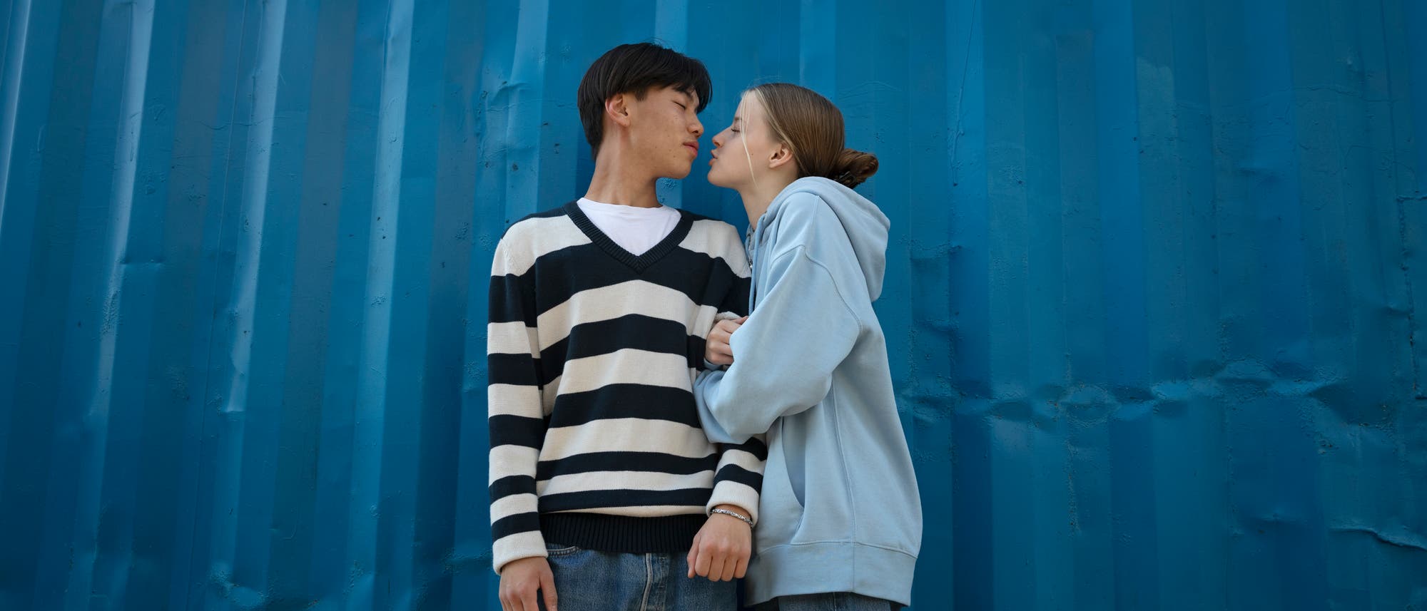 Ein Paar steht vor einer blauen Wand. Die Person links tr&auml;gt einen gestreiften Pullover, die Person rechts eine hellblaue Kapuzenjacke. Sie stehen sich nah gegen&uuml;ber, als ob sie sich gleich k&uuml;ssen w&uuml;rden. Die Szene vermittelt eine intime und liebevolle Stimmung.
