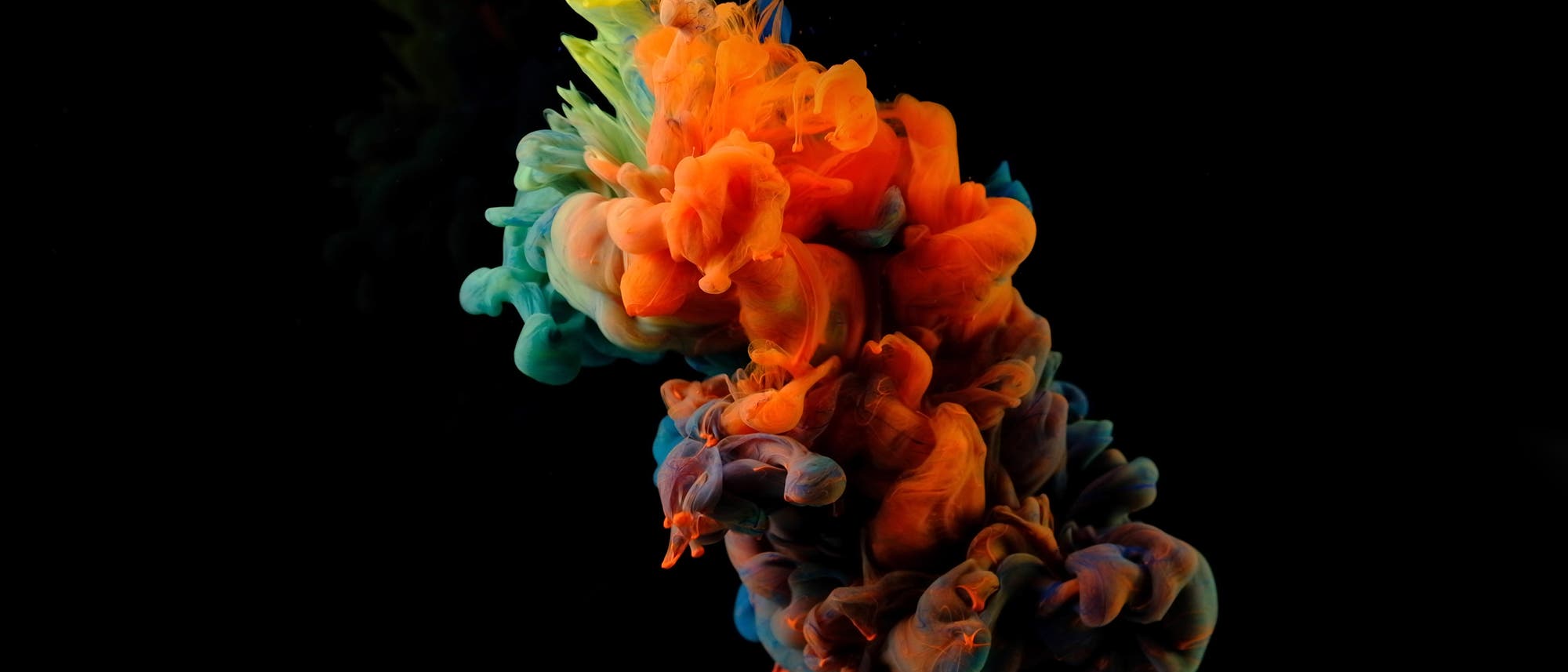 Eine abstrakte Kunstabbildung zeigt leuchtende Farbwolken in Orange, Blau und Grün, die sich in einer fließenden Bewegung vor einem schwarzen Hintergrund vermischen. Die Farben scheinen sich dynamisch zu entfalten und erzeugen einen lebendigen Kontrast zur Dunkelheit des Hintergrunds. Die Komposition vermittelt ein Gefühl von Bewegung und Kreativität.