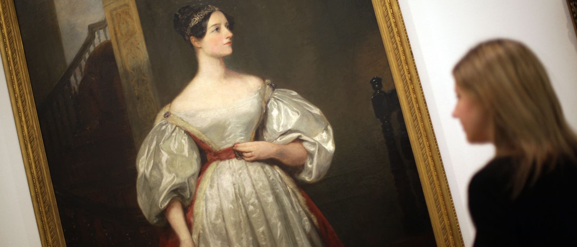 Eine Person betrachtet ein Gemälde in einem Museum. Das Gemälde zeigt eine Frau in einem eleganten, historischen Kleid mit voluminösen Ärmeln und einem roten Band um die Taille. Der Hintergrund des Gemäldes deutet auf eine Treppe hin. Der Rahmen des Gemäldes ist goldfarben. Die Szene vermittelt eine ruhige, nachdenkliche Atmosphäre. Eine Person betrachtet ein Gemälde in einem Museum. Das Gemälde zeigt eine Frau in einem eleganten, historischen Kleid mit voluminösen Ärmeln und einem roten Band um die Taille. Der Hintergrund des Gemäldes deutet auf eine Treppe hin. Der Rahmen des Gemäldes ist goldfarben. Die Szene vermittelt eine ruhige, nachdenkliche Atmosphäre.