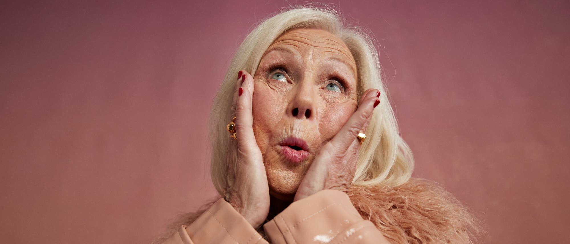 Eine Person mit hellem Haar h&auml;lt ihre H&auml;nde an die Wangen und blickt nach oben, als ob sie &uuml;berrascht oder erfreut ist. Der Hintergrund ist in einem sanften Rosa gehalten. Die Person tr&auml;gt einen beigen Mantel mit einem flauschigen Kragen.