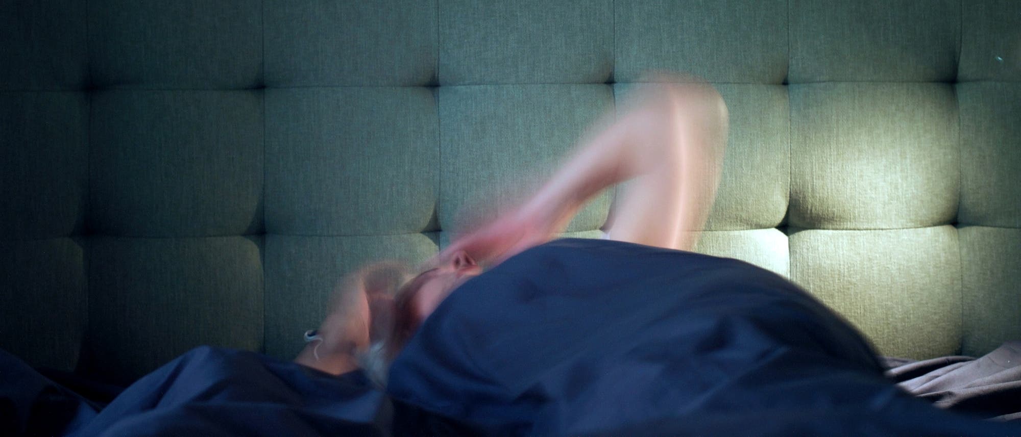 Eine Person liegt in einem Bett mit einem gepolsterten Kopfteil. Die Person ist teilweise von einer dunklen Decke bedeckt und bewegt sich, was durch die verschwommene Darstellung der Arme angedeutet wird. Die Szene ist in ged&auml;mpftem Licht gehalten, was eine ruhige, n&auml;chtliche Atmosph&auml;re schafft.