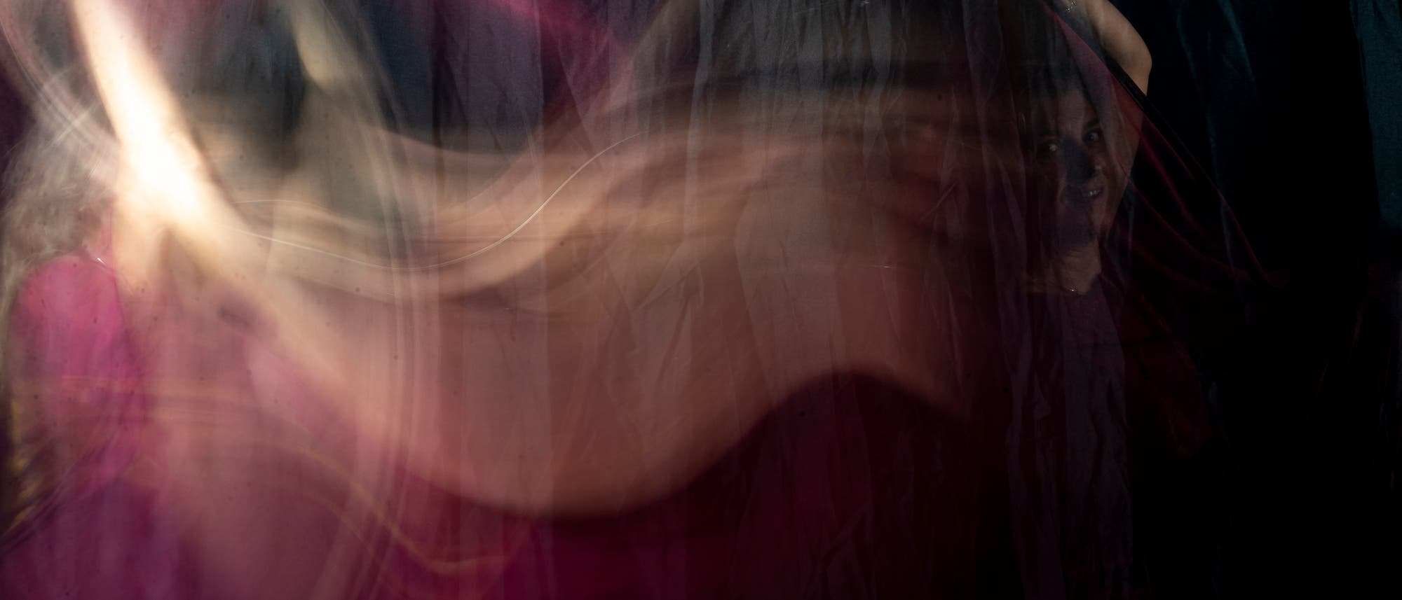 Eine verschwommene, abstrakte Darstellung einer Person, die hinter einem transparenten Vorhang steht. Die Farben sind hauptsächlich dunkle Töne mit rosa und cremefarbenen Akzenten. Die Person hebt einen Arm, während ihr Gesicht teilweise sichtbar ist. Die Szene vermittelt eine geheimnisvolle und künstlerische Atmosphäre. Eine verschwommene, abstrakte Darstellung einer Person, die hinter einem transparenten Vorhang steht. Die Farben sind hauptsächlich dunkle Töne mit rosa und cremefarbenen Akzenten. Die Person hebt einen Arm, während ihr Gesicht teilweise sichtbar ist. Die Szene vermittelt eine geheimnisvolle und künstlerische Atmosphäre.