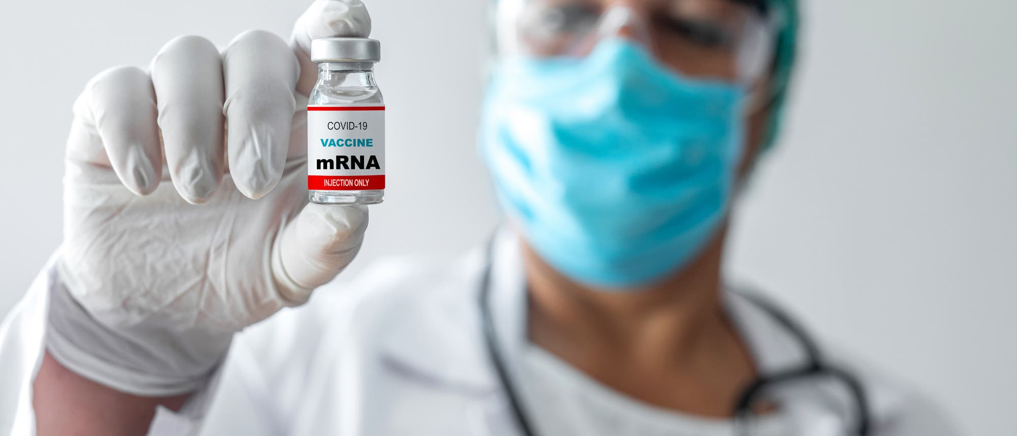 Eine Person in medizinischer Schutzkleidung, einschließlich Maske und Handschuhen, hält ein Fläschchen mit der Aufschrift „COVID-19 Vaccine mRNA“ in die Kamera. Der Hintergrund ist unscharf, der Fokus liegt auf dem Impfstofffläschchen. Eine Person in medizinischer Schutzkleidung, einschließlich Maske und Handschuhen, hält ein Fläschchen mit der Aufschrift „COVID-19 Vaccine mRNA“ in die Kamera. Der Hintergrund ist unscharf, der Fokus liegt auf dem Impfstofffläschchen.