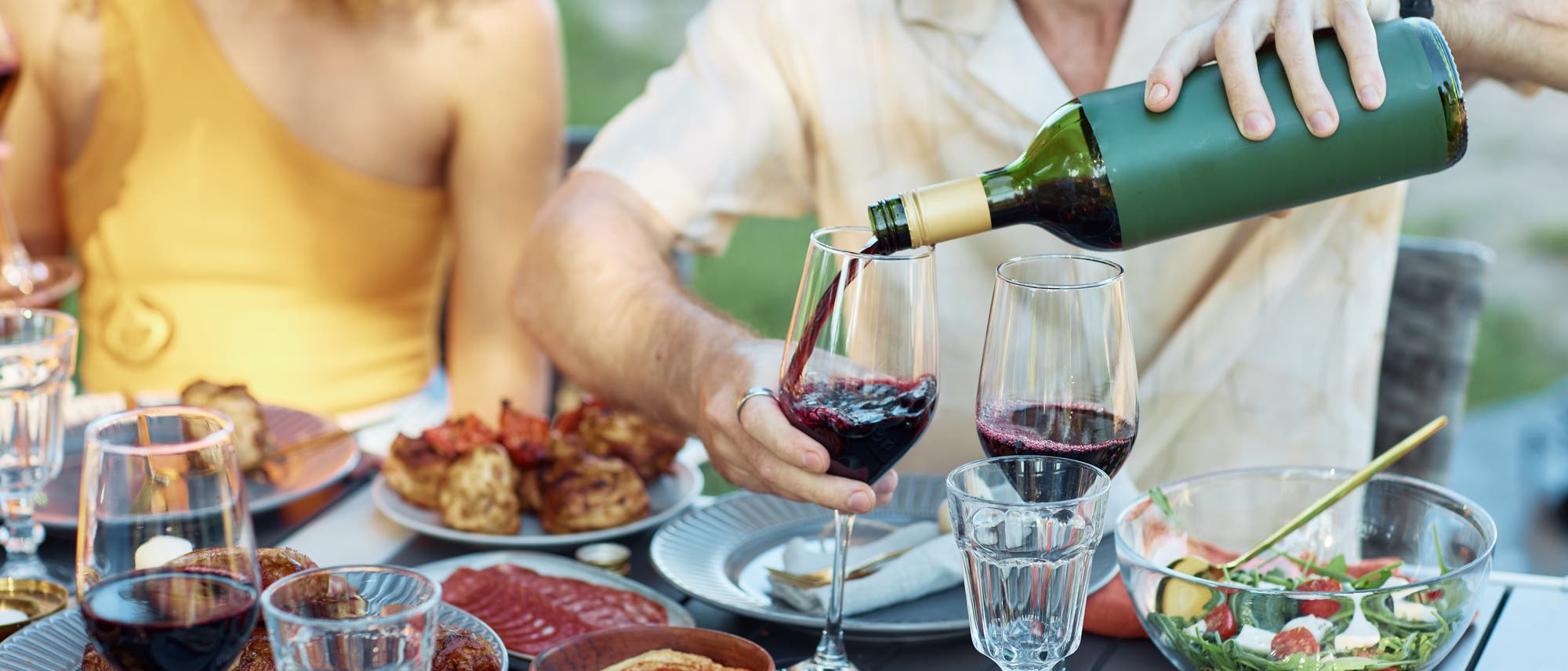 Eine Gruppe von Menschen sitzt an einem Tisch im Freien und genießt ein gemeinsames Essen. Ein Mann schenkt Rotwein in ein Glas ein. Auf dem Tisch stehen verschiedene Speisen wie gegrilltes Fleisch, Salat und Brot.
