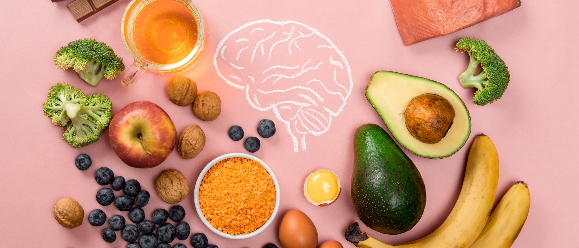Eine Auswahl an Lebensmitteln, die als förderlich für die Gehirngesundheit gelten, auf einem rosa Hintergrund. Zu sehen sind Brokkoli, ein Apfel, Walnüsse, Blaubeeren, Linsen, Eier, Bananen, Avocado, Lachs, Schokolade und ein Glas Tee. In der Mitte ist eine stilisierte Zeichnung eines Gehirns zu erkennen. Eine Auswahl an Lebensmitteln, die als förderlich für die Gehirngesundheit gelten, auf einem rosa Hintergrund. Zu sehen sind Brokkoli, ein Apfel, Walnüsse, Blaubeeren, Linsen, Eier, Bananen, Avocado, Lachs, Schokolade und ein Glas Tee. In der Mitte ist eine stilisierte Zeichnung eines Gehirns zu erkennen.