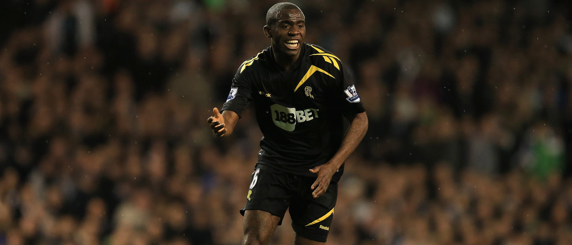 Fabrice Muamba in einem schwarzen Trikot läuft auf einem Spielfeld. Der Spieler scheint in Aktion zu sein, mit einem entschlossenen Ausdruck. Im Hintergrund sind unscharfe Zuschauer auf den Tribünen zu erkennen. Fabrice Muamba in einem schwarzen Trikot läuft auf einem Spielfeld. Der Spieler scheint in Aktion zu sein, mit einem entschlossenen Ausdruck. Im Hintergrund sind unscharfe Zuschauer auf den Tribünen zu erkennen.