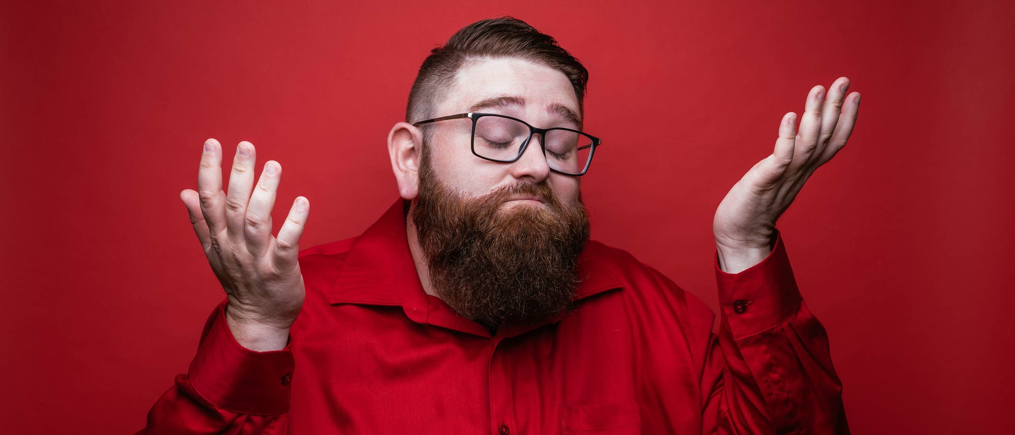 Ein Mann mit Brille und Bart trägt ein rotes Hemd und hebt beide Hände mit einem etwas übertrieben wirkenden Gesichtsausdruck, als ob er ratlos sei. Der Hintergrund ist ebenfalls rot. Ein Mann mit Brille und Bart trägt ein rotes Hemd und hebt beide Hände mit einem etwas übertrieben wirkenden Gesichtsausdruck, als ob er ratlos sei. Der Hintergrund ist ebenfalls rot.
