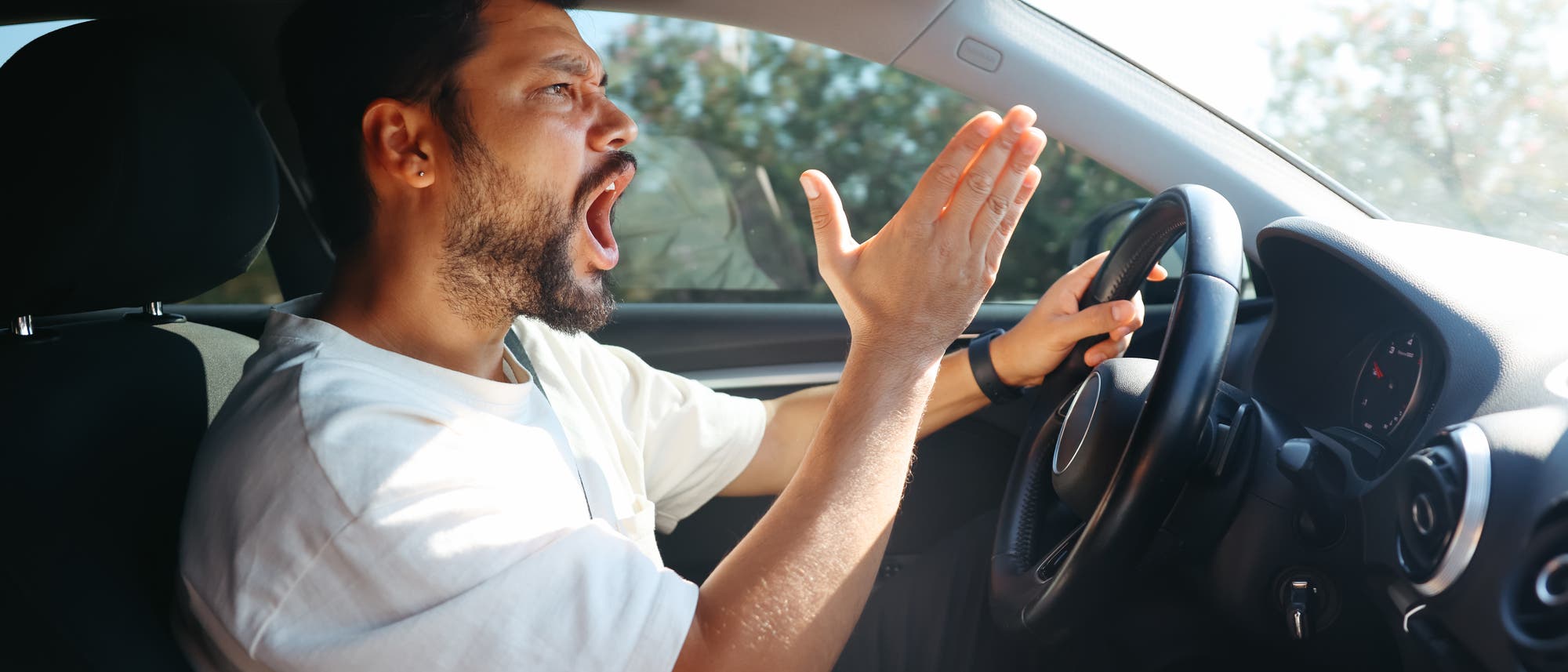 Ein Mann sitzt am Steuer eines Autos und zeigt eine frustrierte oder verärgerte Geste mit der rechten Hand erhoben. Er trägt ein weißes T-Shirt und hat dunkles Haar und einen Bart. Die Sonne scheint durch das Autofenster, und im Hintergrund sind verschwommene Bäume zu sehen. Ein Mann sitzt am Steuer eines Autos und zeigt eine frustrierte oder verärgerte Geste mit der rechten Hand erhoben. Er trägt ein weißes T-Shirt und hat dunkles Haar und einen Bart. Die Sonne scheint durch das Autofenster, und im Hintergrund sind verschwommene Bäume zu sehen.