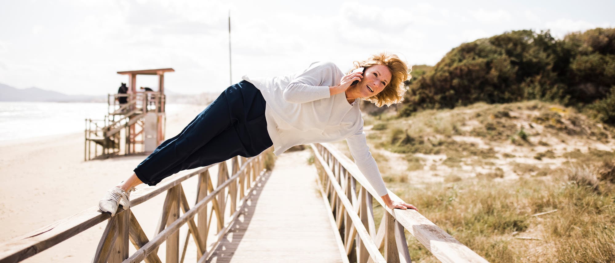 Eine Person in sportlicher Kleidung balanciert seitlich auf einem Holzgeländer einer Strandpromenade und telefoniert dabei. Im Hintergrund sind ein Rettungsturm, das Meer und Dünen zu sehen. Der Himmel ist bewölkt, die Sonne scheint.
