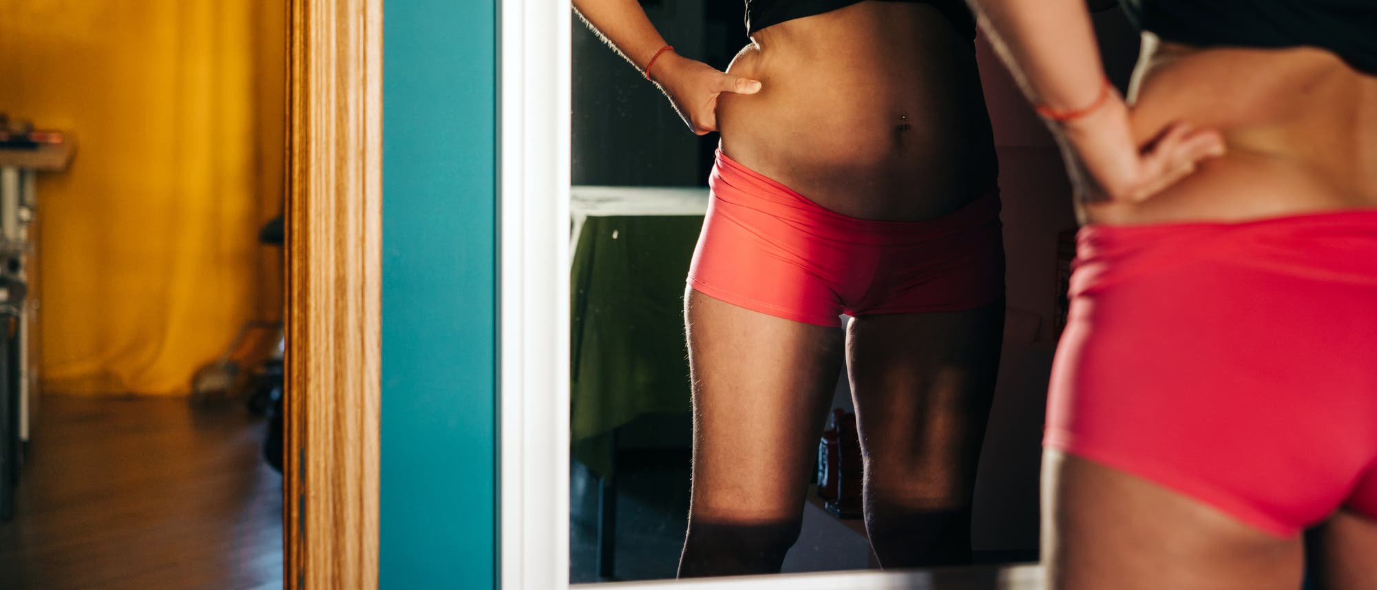 Eine Person in Sportkleidung betrachtet sich in einem Spiegel. Die Szene zeigt einen Raum mit einem blauen und gelben Farbschema. Die Person steht mit den H&auml;nden in die H&uuml;ften gest&uuml;tzt vor dem Spiegel.