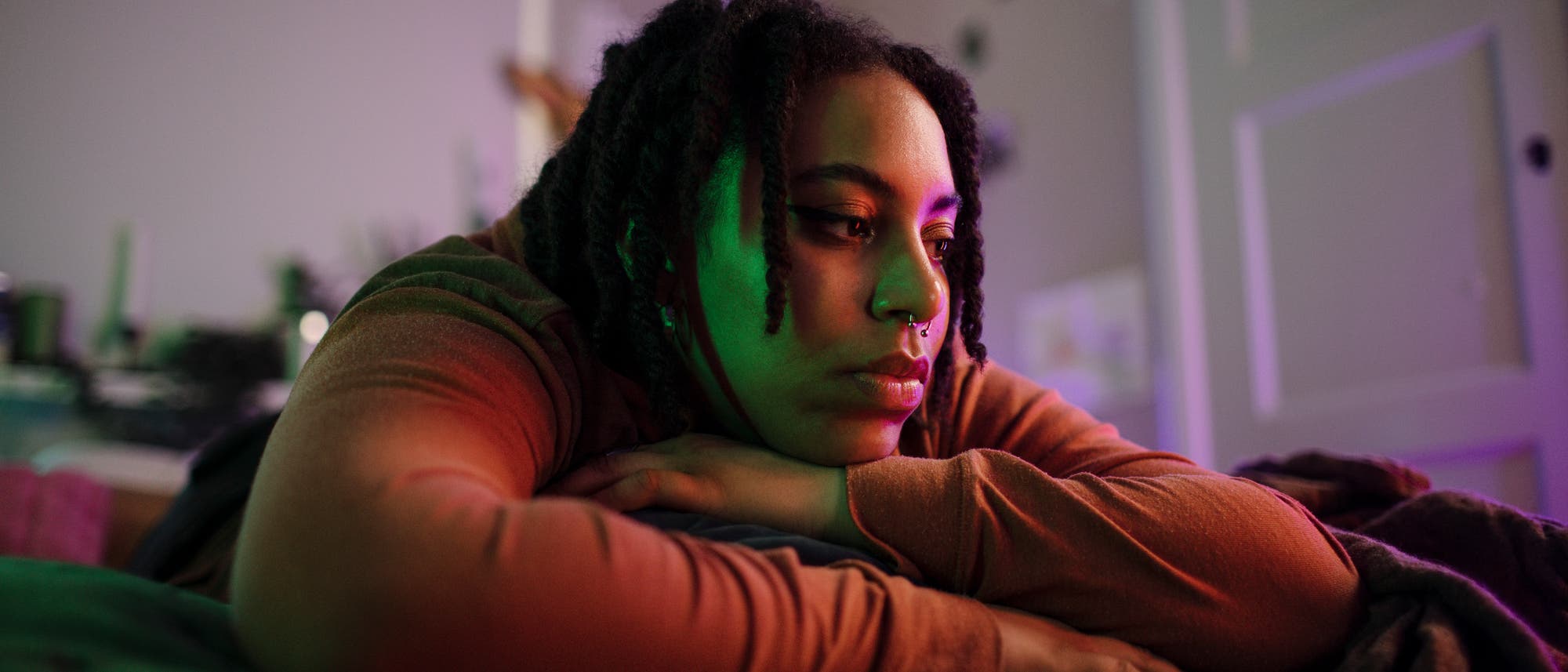 Eine Person liegt nachdenklich auf einem Bett, den Kopf auf die verschränkten Arme gestützt. Der Raum ist in sanftes, farbiges Licht getaucht, das eine ruhige und nachdenkliche Atmosphäre schafft. Im Hintergrund sind unscharfe Details eines Zimmers zu erkennen.