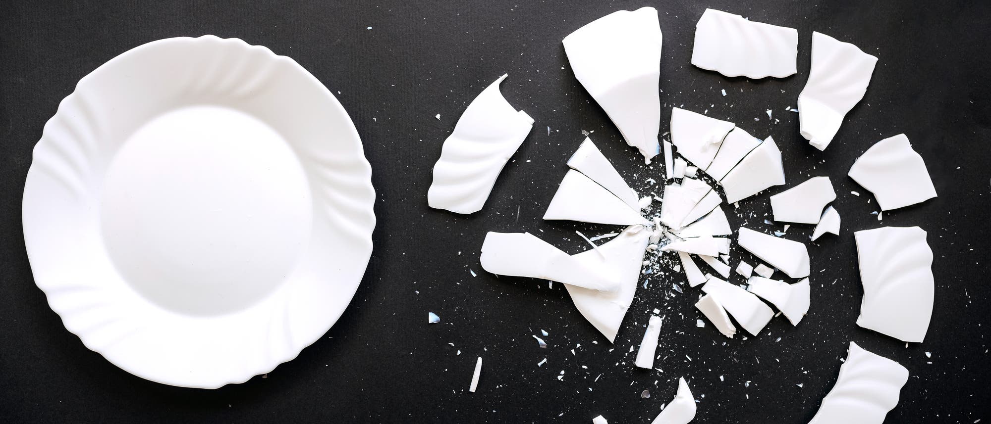 Ein unversehrter weißer Teller liegt auf einer dunklen Oberfläche, während daneben die Scherben eines identischen zerbrochenen Tellers verstreut sind. Ein unversehrter weißer Teller liegt auf einer dunklen Oberfläche, während daneben die Scherben eines identischen zerbrochenen Tellers verstreut sind.