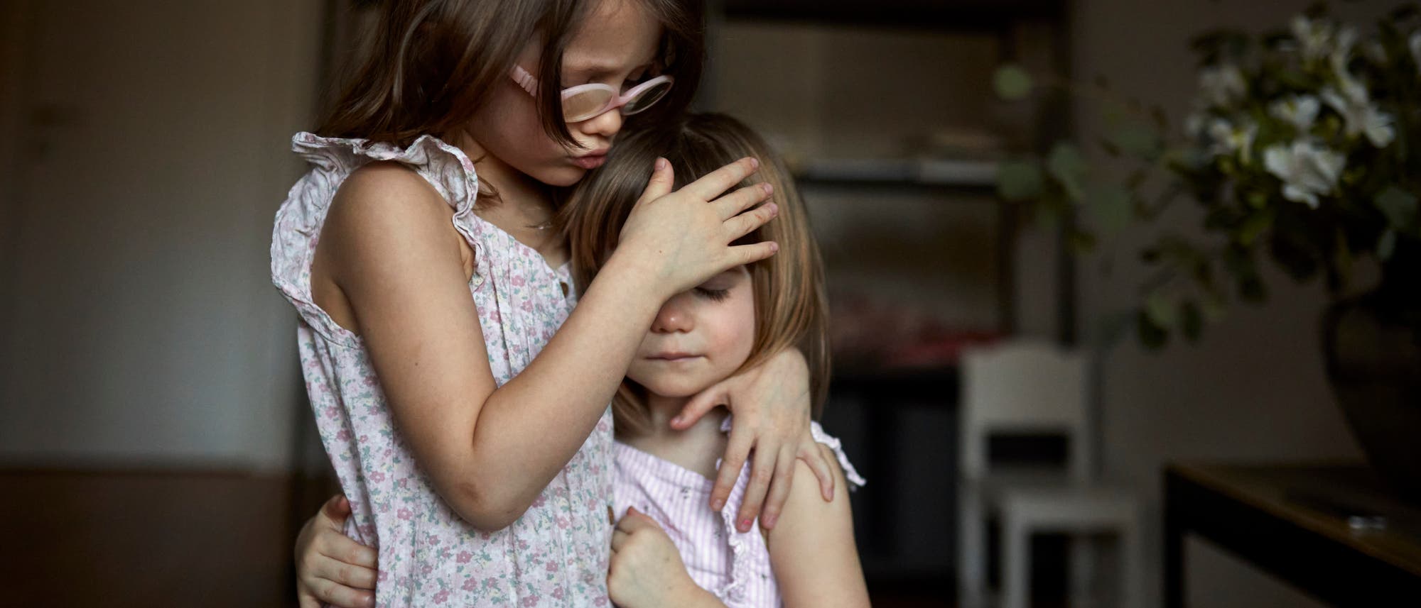 Zwei Kinder in sommerlichen Kleidern stehen in einem Raum. Das ältere Kind umarmt das jüngere und legt tröstend eine Hand auf dessen Kopf. Beide haben die Augen geschlossen. Im Hintergrund sind ein Regal und ein Tisch mit Blumen zu sehen. Die Szene vermittelt ein Gefühl von Geborgenheit und Zuneigung.