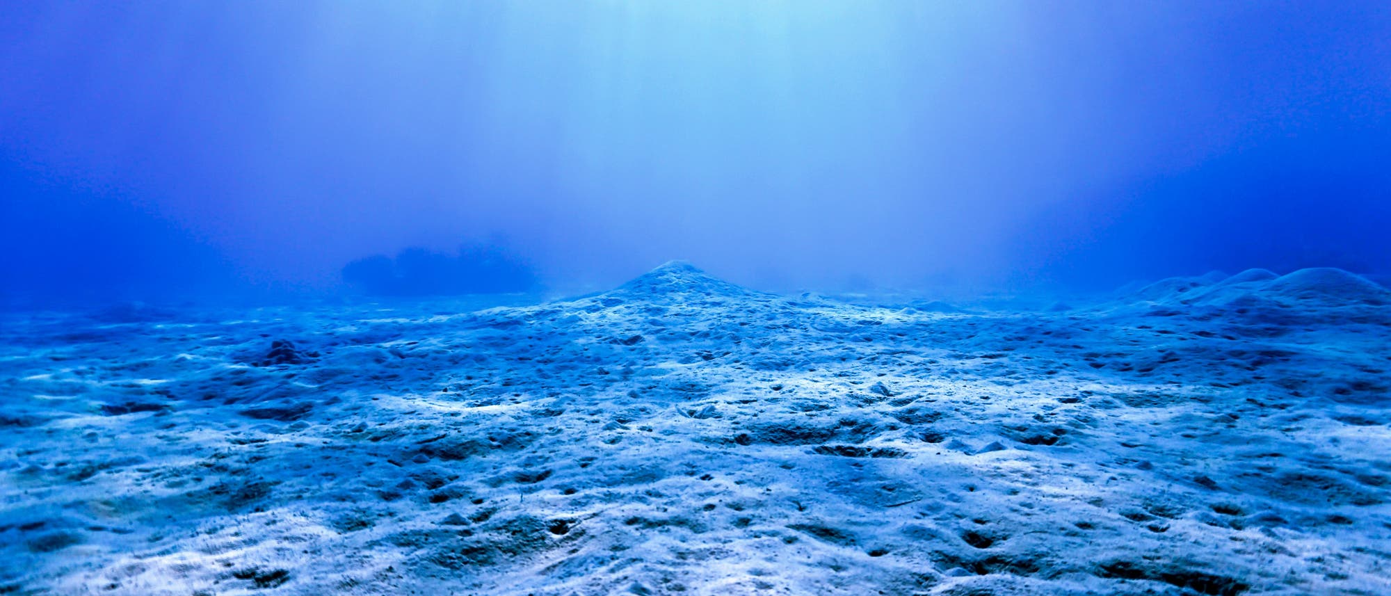 Unterwasseraufnahme eines Meeresbodens, der von Sonnenstrahlen beleuchtet wird. Die sandige Oberfläche ist uneben und erstreckt sich bis zum Horizont, wo das Wasser in ein tiefes Blau übergeht. Die Lichtstrahlen erzeugen ein sanftes, diffuses Leuchten im Wasser.