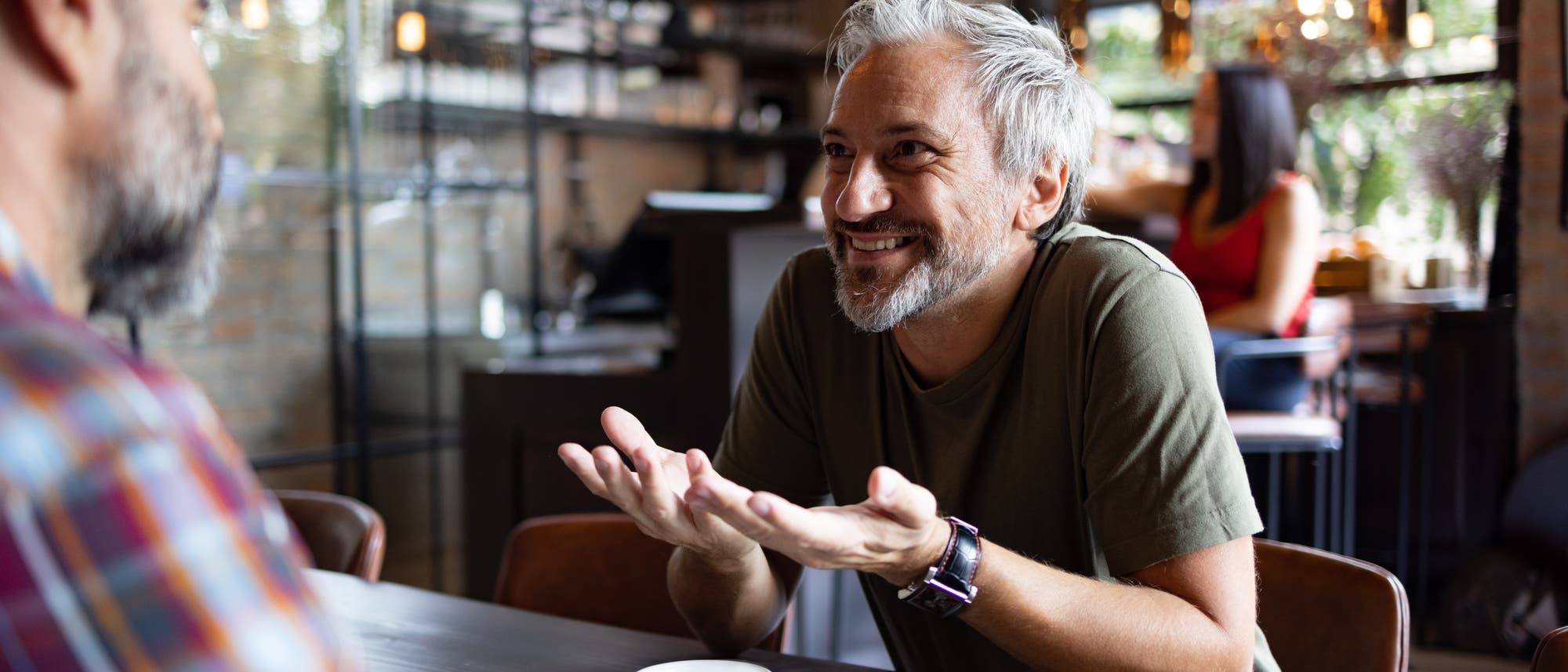 Ein Mann mit grauen Haaren und Bart sitzt in einem Café und unterhält sich lebhaft mit einem anderen Mann, der ihm gegenübersitzt. Auf dem Tisch stehen Kaffeetassen und Gläser mit Wasser. Im Hintergrund ist eine Frau zu sehen, die an einem Tisch sitzt. Die Atmosphäre ist gemütlich und einladend, mit warmem Licht und moderner Einrichtung. Ein Mann mit grauen Haaren und Bart sitzt in einem Café und unterhält sich lebhaft mit einem anderen Mann, der ihm gegenübersitzt. Auf dem Tisch stehen Kaffeetassen und Gläser mit Wasser. Im Hintergrund ist eine Frau zu sehen, die an einem Tisch sitzt. Die Atmosphäre ist gemütlich und einladend, mit warmem Licht und moderner Einrichtung.