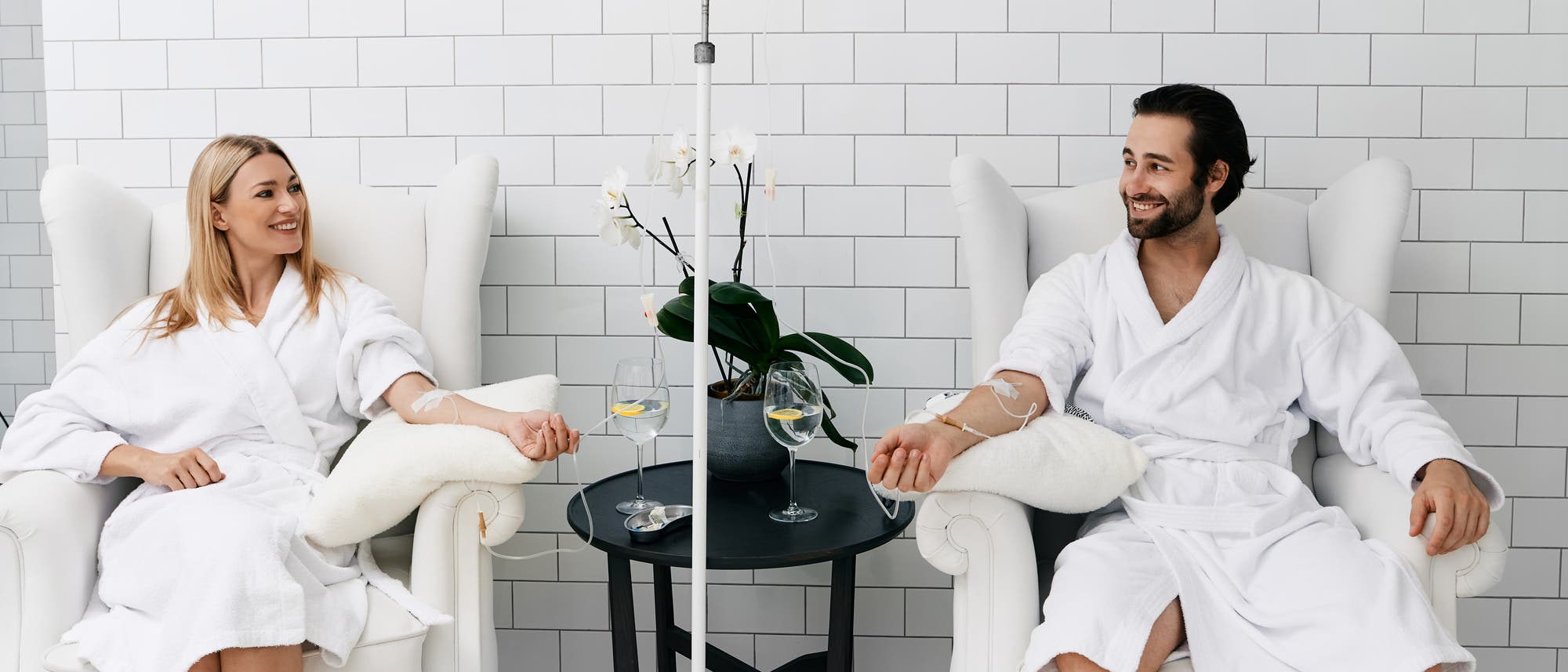 Zwei Personen in weißen Bademänteln sitzen in bequemen Sesseln in einem hellen Raum mit weißen Fliesenwänden. Beide erhalten eine Infusion über einen intravenösen Zugang am Arm. Zwischen ihnen steht ein kleiner Tisch mit einer Orchidee und zwei Gläsern Wasser. Sie lächeln und wirken entspannt.