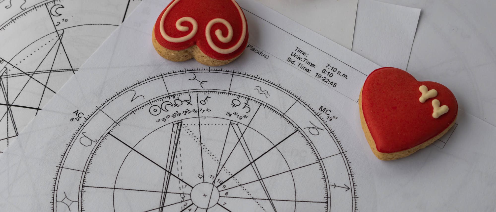 Ein Bild zeigt astrologische Diagramme mit verschiedenen Symbolen und Linien, die auf Papier gedruckt sind. Auf den Diagrammen liegen herzförmige Kekse mit rotem Zuckerguss und dekorativen Mustern. Die Kekse sind in verschiedenen Designs gestaltet, darunter Spiralen und kleine Herzen. Das Bild kombiniert wissenschaftliche und dekorative Elemente und vermittelt eine Mischung aus Astrologie und kulinarischer Kunst.