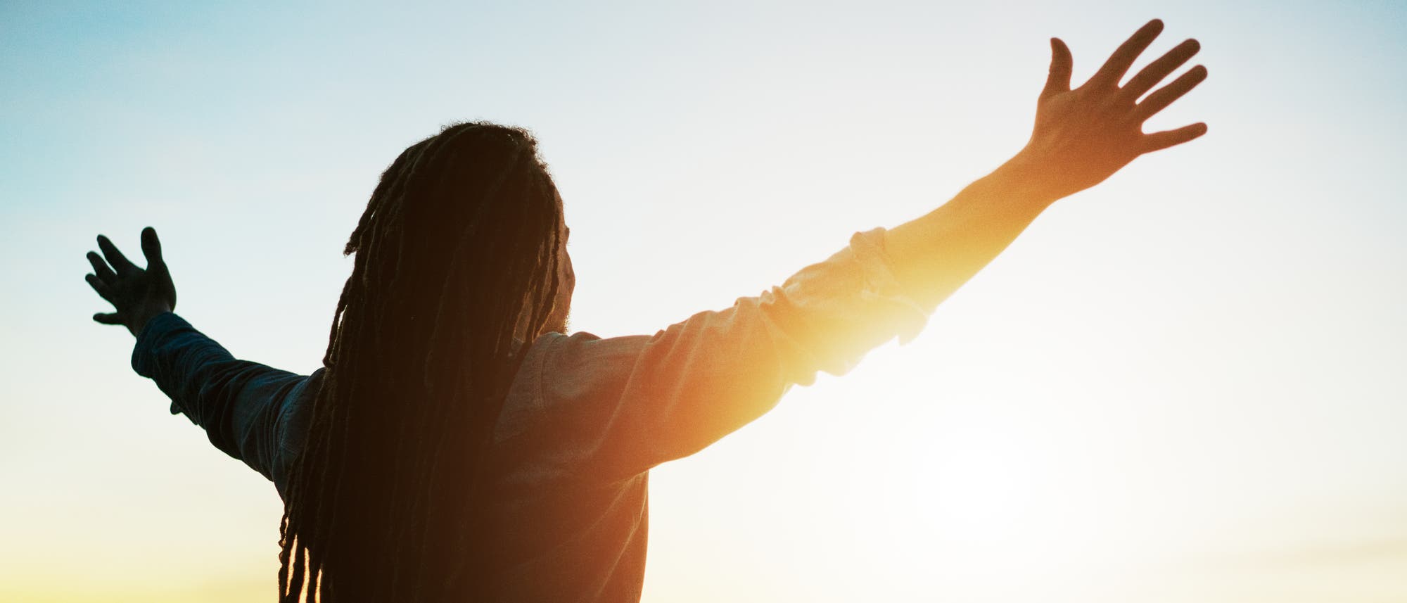 Eine Person steht mit ausgebreiteten Armen im Freien und blickt in einen farbenfrohen Sonnenuntergang. Der Himmel zeigt warme T&ouml;ne von Orange und Blau. Die Szene vermittelt ein Gef&uuml;hl von Freiheit und Freude.