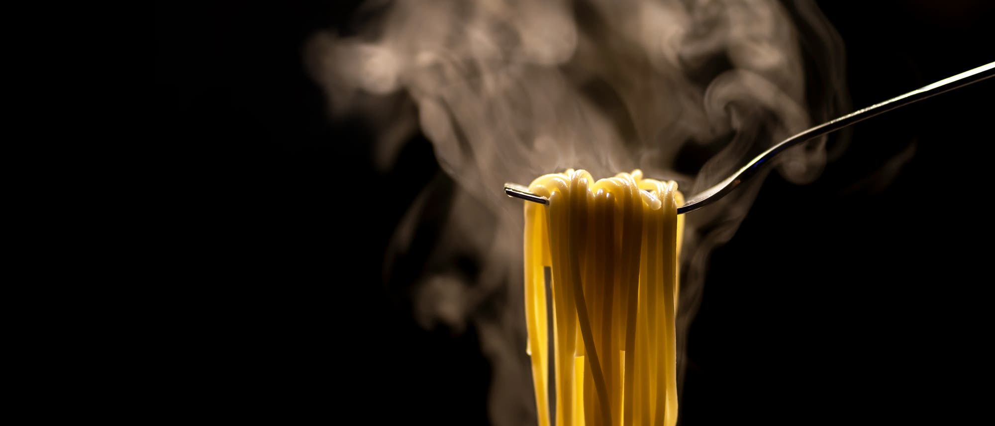 Eine Gabel h&auml;lt dampfende Spaghetti vor einem dunklen Hintergrund. Der Dampf steigt in weichen Schwaden auf, was auf die Frische und W&auml;rme der Pasta hinweist. Die Beleuchtung hebt die goldene Farbe der Spaghetti hervor.