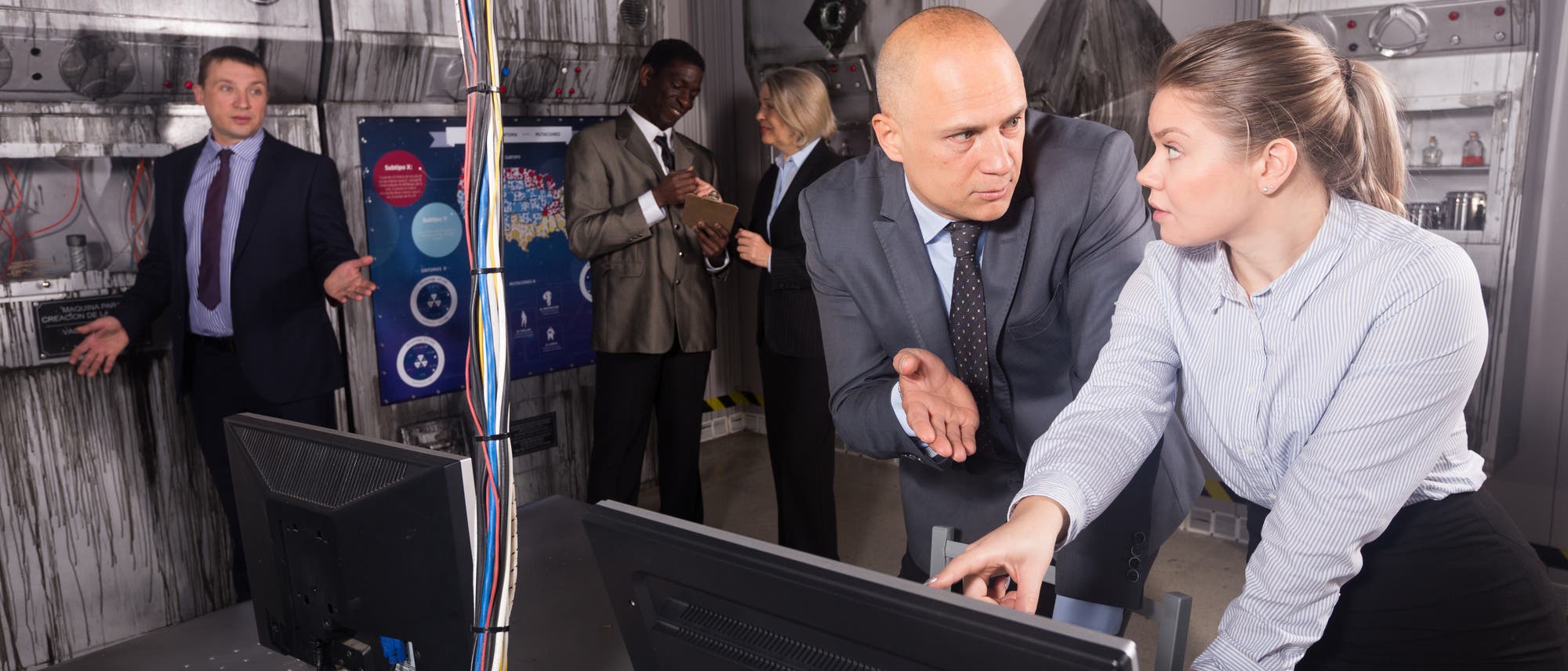 Eine Gruppe von Personen in einem Raum, der wie ein Kontrollzentrum aussieht. Zwei Personen im Vordergrund diskutieren angeregt über einen Computerbildschirm. Im Hintergrund sind weitere Personen zu sehen, die miteinander sprechen und Notizen machen. An den Wänden befinden sich technische Geräte und ein Poster mit Diagrammen. Die Szene vermittelt eine geschäftige und konzentrierte Arbeitsatmosphäre.