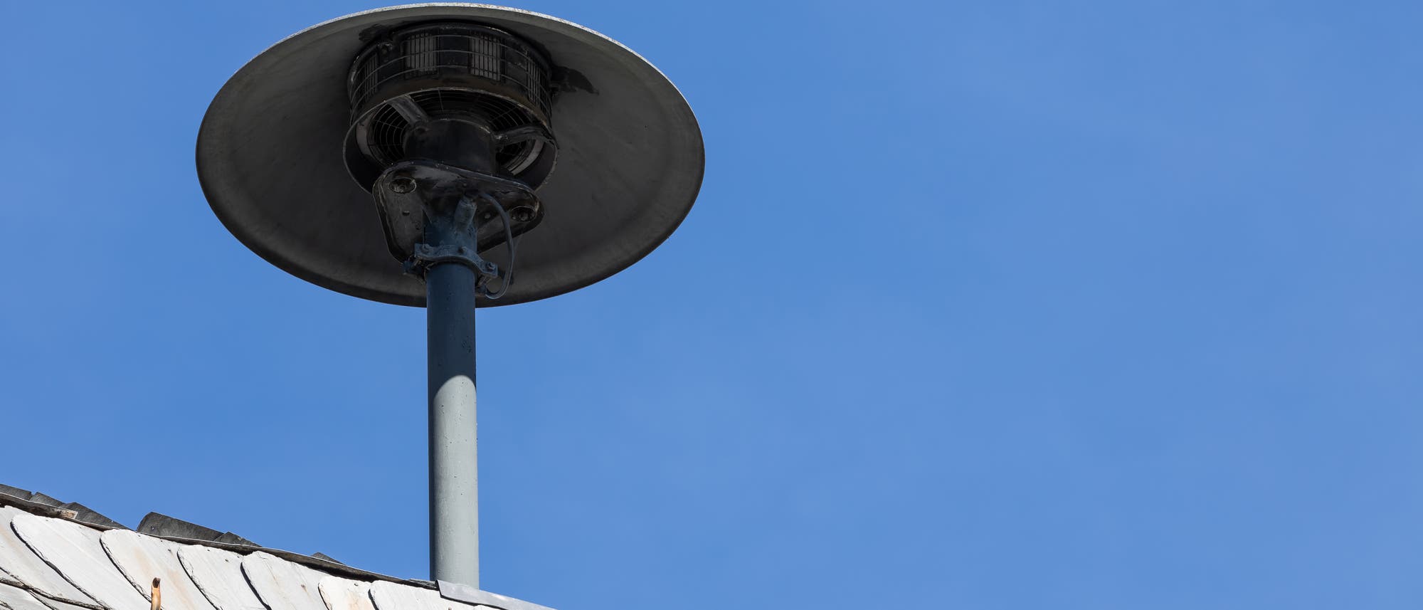 Ein grauer Sirenenmast ragt über ein mit Schieferplatten gedecktes Dach in den blauen Himmel. Die Sirene hat eine runde, schalenförmige Abdeckung. Das Bild zeigt eine klare, wolkenlose Wetterlage.