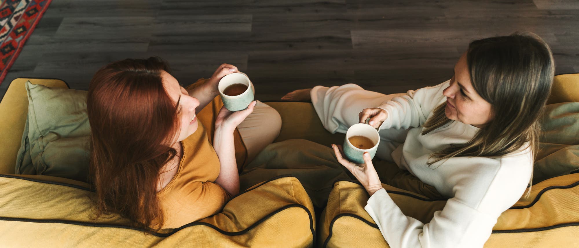 Zwei Personen sitzen auf einem gelben Sofa und halten Tassen mit einem Hei&szlig;getr&auml;nk. Sie unterhalten sich in einer gem&uuml;tlichen Wohnumgebung mit Holzboden und einem bunten Teppich im Hintergrund. Die Szene vermittelt eine entspannte und freundliche Atmosph&auml;re.