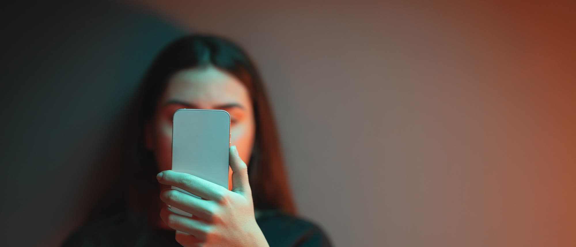 Mädchen um die 18 Jahre starrt auf ihr Handy, das sie vor dem Gesicht hält. Sie steht vor einer weißen Wand, die leicht orange beleuchtet ist.  