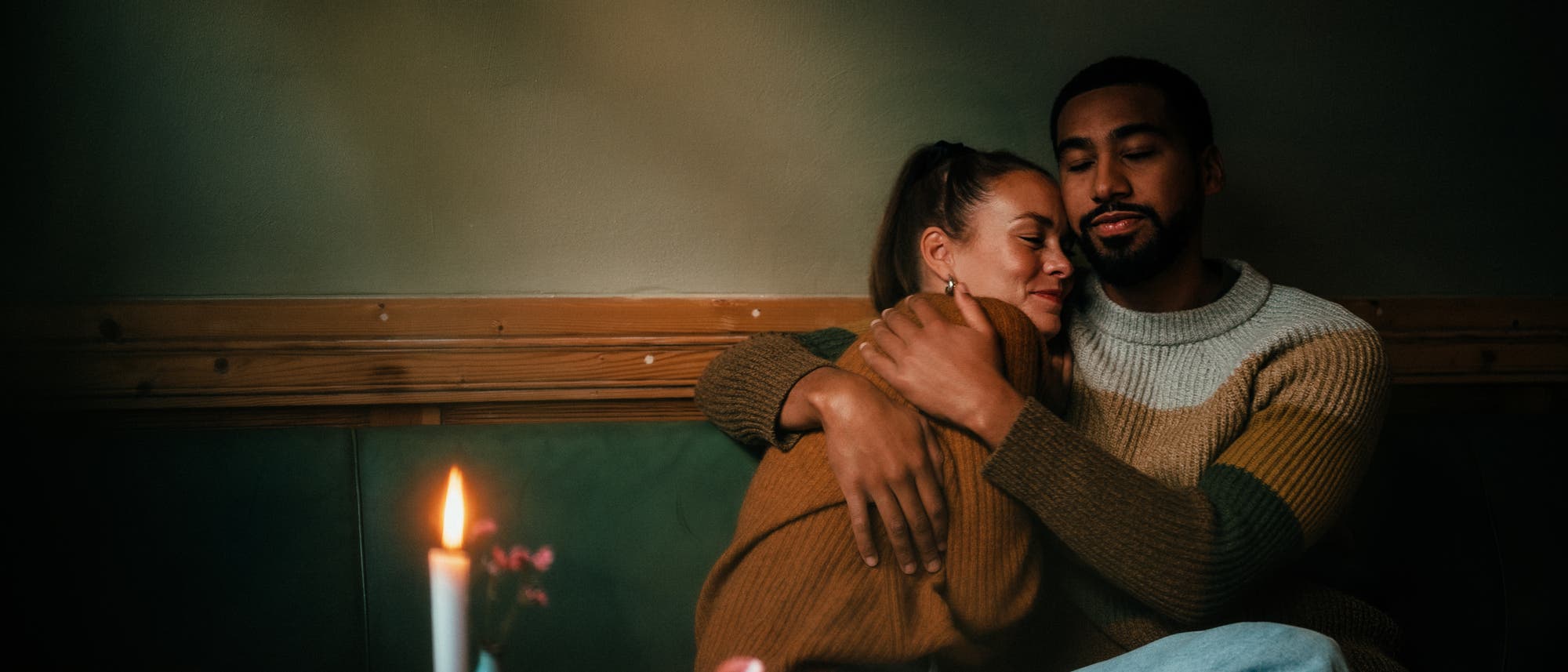 Ein Paar sitzt in einem gem&uuml;tlichen Caf&eacute; und umarmt sich liebevoll. Auf dem Tisch vor ihnen stehen zwei Tassen Kaffee, eine brennende Kerze und ein kleines Geschenk mit rotem Band. Die Atmosph&auml;re ist warm und intim, mit ged&auml;mpftem Licht und einem Gef&uuml;hl von Geborgenheit.