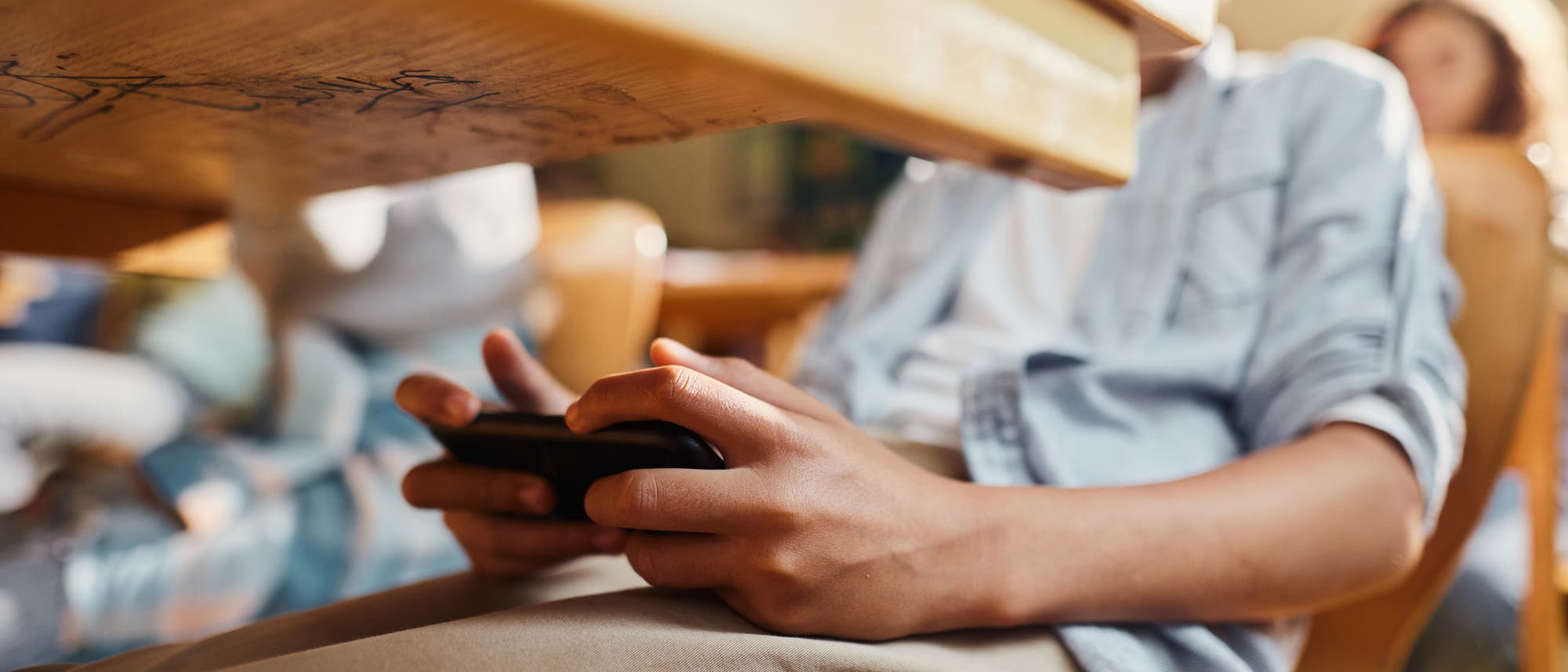 Ein Kind sitzt in einem Klassenzimmer und hält ein Smartphone unter dem Tisch. Der Fokus liegt auf den Händen und dem Gerät. Im Hintergrund sind verschwommen andere Schüler zu sehen. Die Szene vermittelt eine alltägliche Schulsituation.