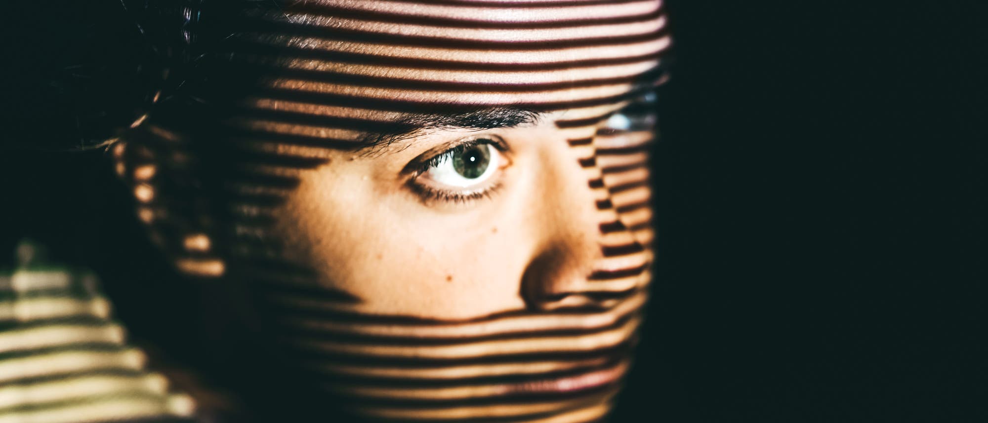 Eine Person schaut nach rechts, während horizontale Lichtstreifen ihr Gesicht überqueren und einen dramatischen Schatteneffekt erzeugen. Der Hintergrund ist dunkel, wodurch das Gesicht und die Lichtmuster hervorgehoben werden. Eine Person schaut nach rechts, während horizontale Lichtstreifen ihr Gesicht überqueren und einen dramatischen Schatteneffekt erzeugen. Der Hintergrund ist dunkel, wodurch das Gesicht und die Lichtmuster hervorgehoben werden.