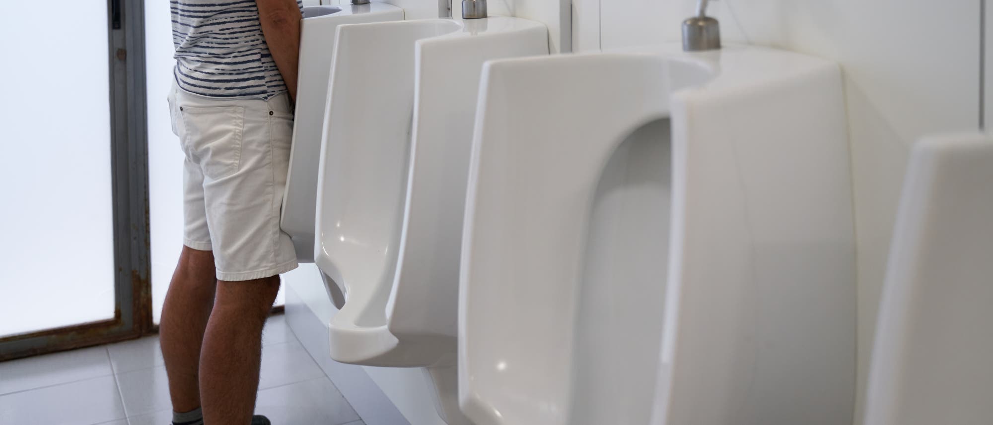 Eine Person steht in einem &ouml;ffentlichen Badezimmer vor einer Reihe von Urinalen. Die Person tr&auml;gt ein gestreiftes T-Shirt, wei&szlig;e Shorts und dunkle Schuhe. Die Umgebung ist sauber und modern gestaltet.