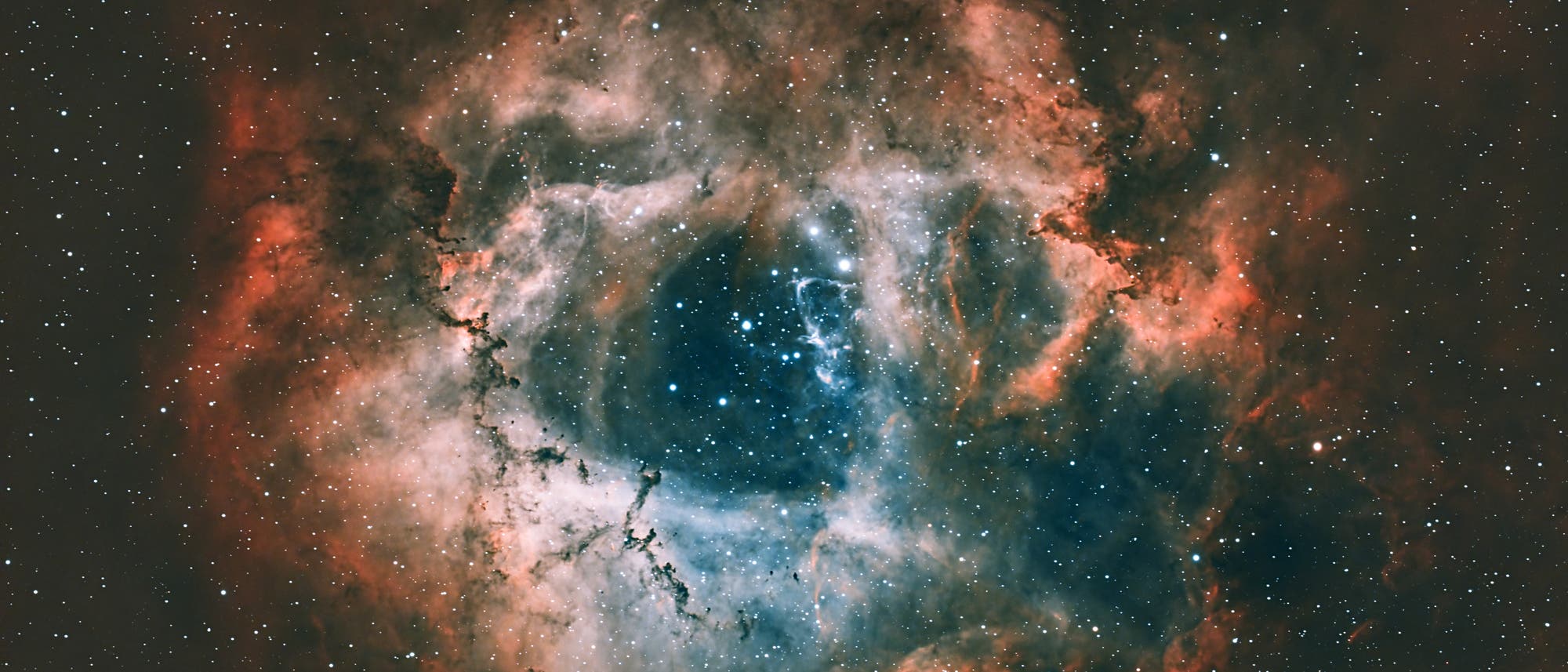 Ein farbenpr&auml;chtiges astronomisches Bild zeigt einen Nebel im Weltraum. Die zentrale Region ist dunkelblau und von leuchtend roten und orangefarbenen Gaswolken umgeben. Zahlreiche Sterne sind im gesamten Bild sichtbar, die in der Dunkelheit des Universums funkeln. Der Nebel hat eine blumenartige Form, die an eine Rosette erinnert. Dieses Bild veranschaulicht die Sch&ouml;nheit und Komplexit&auml;t kosmischer Strukturen.