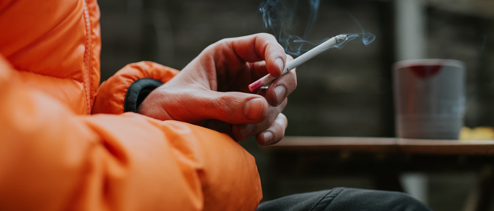 Eine Person in einer orangefarbenen Jacke h&auml;lt eine brennende Zigarette in der Hand. Rauch steigt von der Zigarette auf. Im Hintergrund ist ein unscharfer Tisch mit einem Becher zu sehen. Die Szene spielt sich im Freien ab.