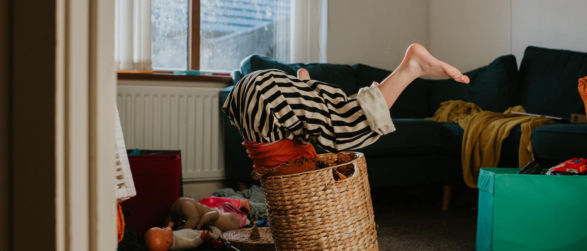 Ein Kind in gestreifter Hose steckt kopf&uuml;ber in einem geflochtenen Korb in einem Wohnzimmer. Im Hintergrund sind ein Sofa, ein Fenster und verstreutes Spielzeug zu sehen. Die Szene wirkt verspielt und chaotisch.