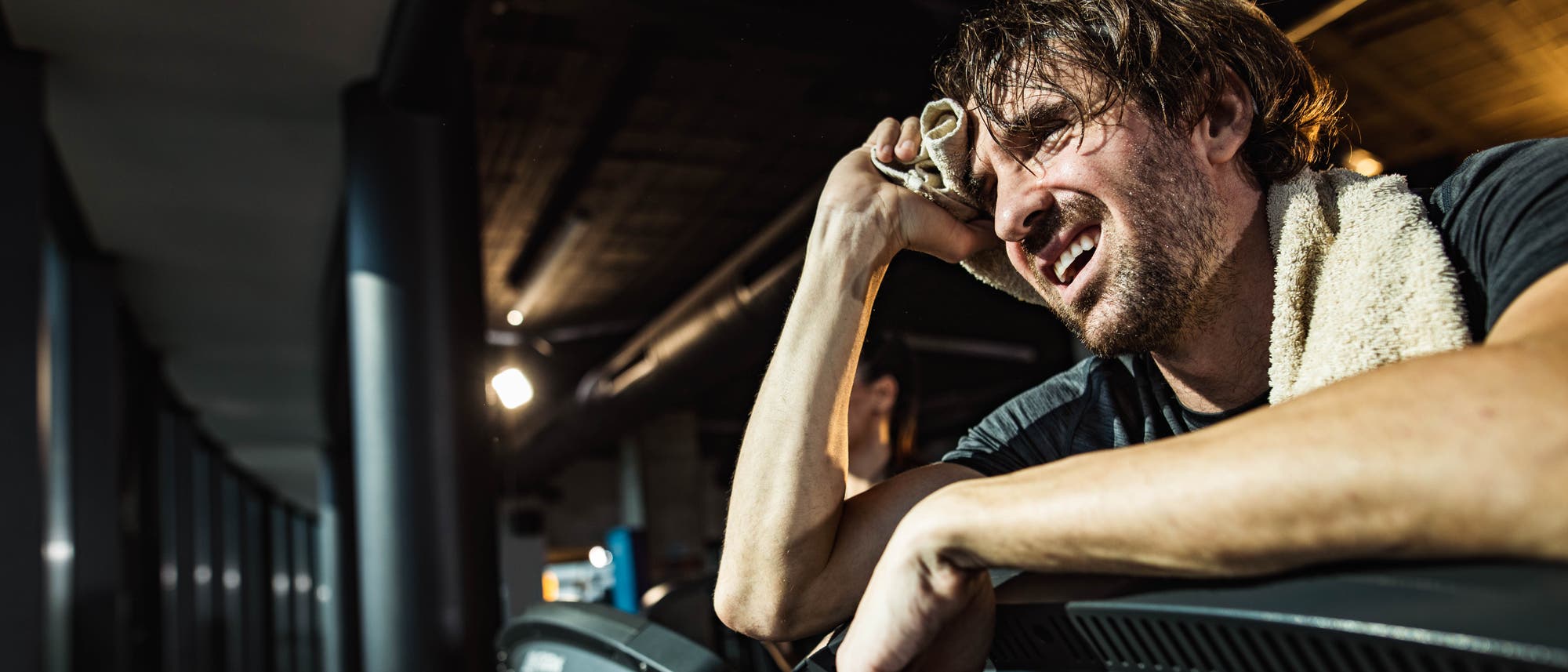 Ein Mann im Fitnessstudio lehnt erschöpft auf einem Laufband oder Fahrradergonometer nach dem Training. Er hält ein Handtuch an die Stirn und hat ein weiteres Handtuch um den Hals. Im Hintergrund sind weitere Fitnessgeräte zu sehen. Ein Mann im Fitnessstudio lehnt erschöpft auf einem Laufband oder Fahrradergonometer nach dem Training. Er hält ein Handtuch an die Stirn und hat ein weiteres Handtuch um den Hals. Im Hintergrund sind weitere Fitnessgeräte zu sehen.