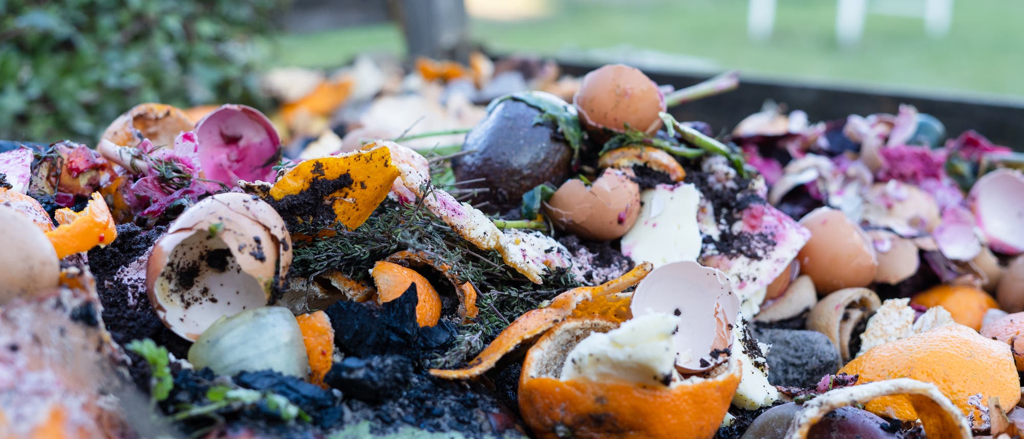 Ein Komposthaufen im Freien mit verschiedenen organischen Abf&auml;llen wie Eierschalen, Obst- und Gem&uuml;seschalen sowie Pflanzenresten. Im Hintergrund ist ein unscharfer Gartenbereich zu sehen. Der Komposthaufen zeigt den Prozess der nat&uuml;rlichen Zersetzung und das Recycling von K&uuml;chenabf&auml;llen.