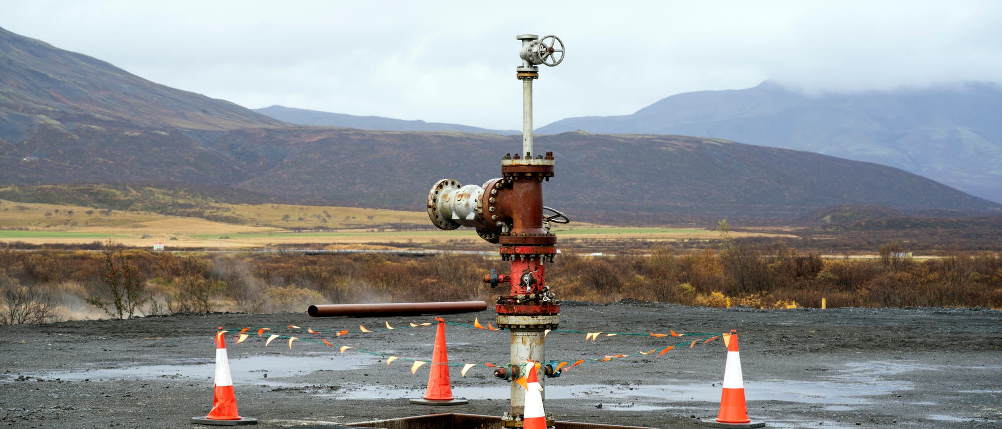 Ein einzelnes Bohrlochventil steht auf einer asphaltierten Fl&auml;che, umgeben von orange-wei&szlig;en Verkehrskegeln und Absperrband. Im Hintergrund erstreckt sich eine h&uuml;gelige Landschaft mit sp&auml;rlicher Vegetation unter einem bew&ouml;lkten Himmel.