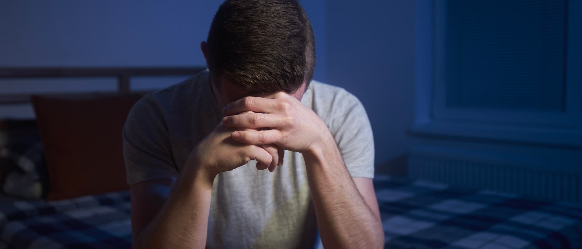 Eine Person sitzt auf einem Bett in einem abgedunkelten Raum, den Kopf in die Hände gestützt. Die Atmosphäre wirkt nachdenklich und melancholisch. Im Hintergrund ist ein Fenster mit geschlossenen Jalousien zu sehen. Eine Person sitzt auf einem Bett in einem abgedunkelten Raum, den Kopf in die Hände gestützt. Die Atmosphäre wirkt nachdenklich und melancholisch. Im Hintergrund ist ein Fenster mit geschlossenen Jalousien zu sehen.